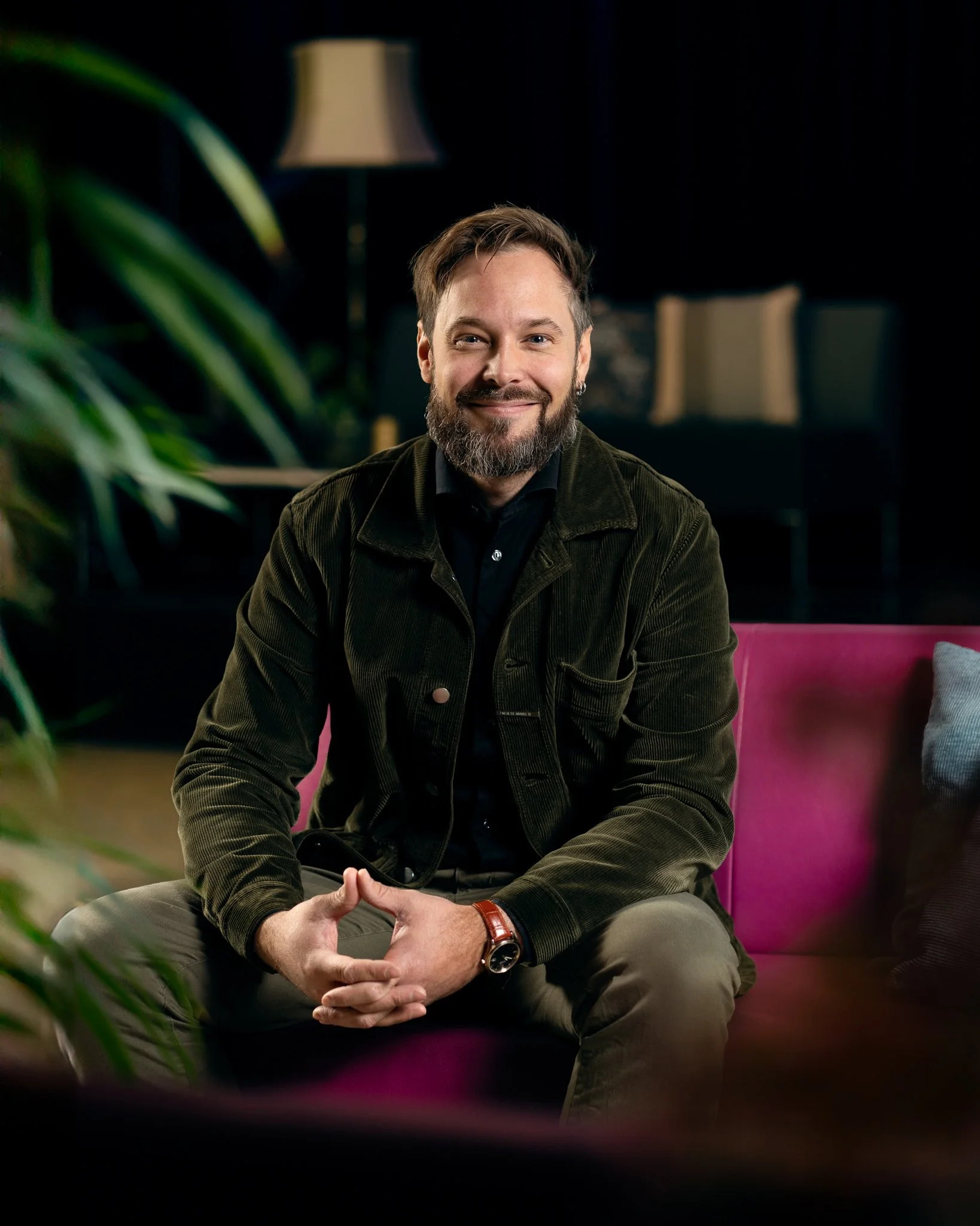 En smilende mann med skjegg, sittende på en rosa sofa i et rom med mørke vegger, green plants, og et bord med en lampskulptur i bakgrunnen.