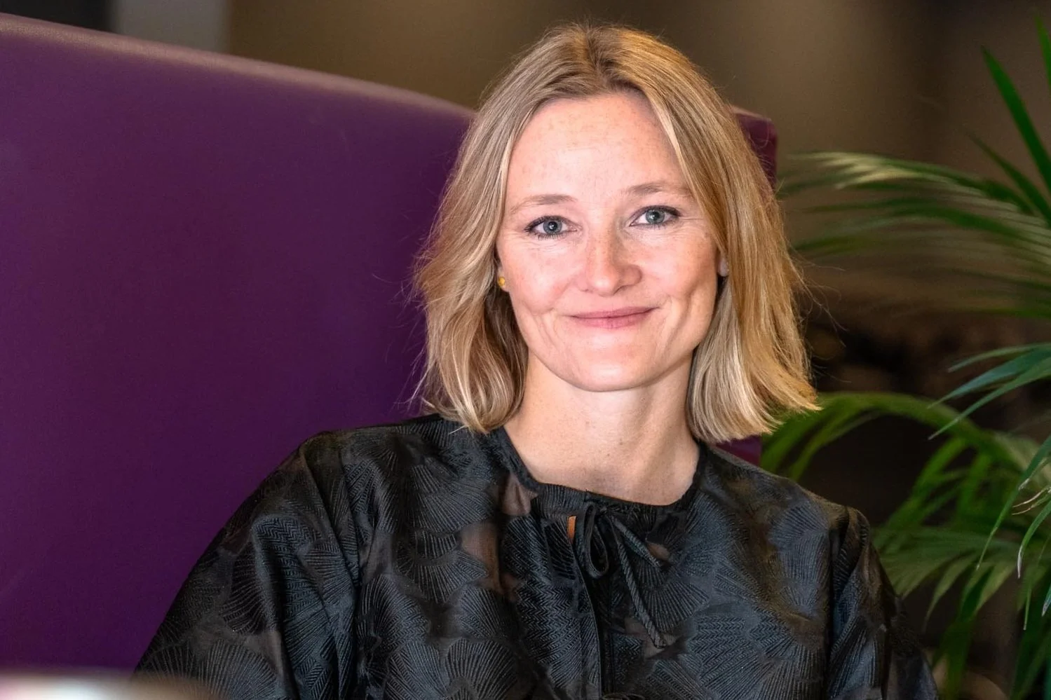 Et portrett av en smilende kvinne med mellomlang blond hår, kledd i svart bluse, sittende mot en lilla vegg med grønt planteliv i bakgrunnen.