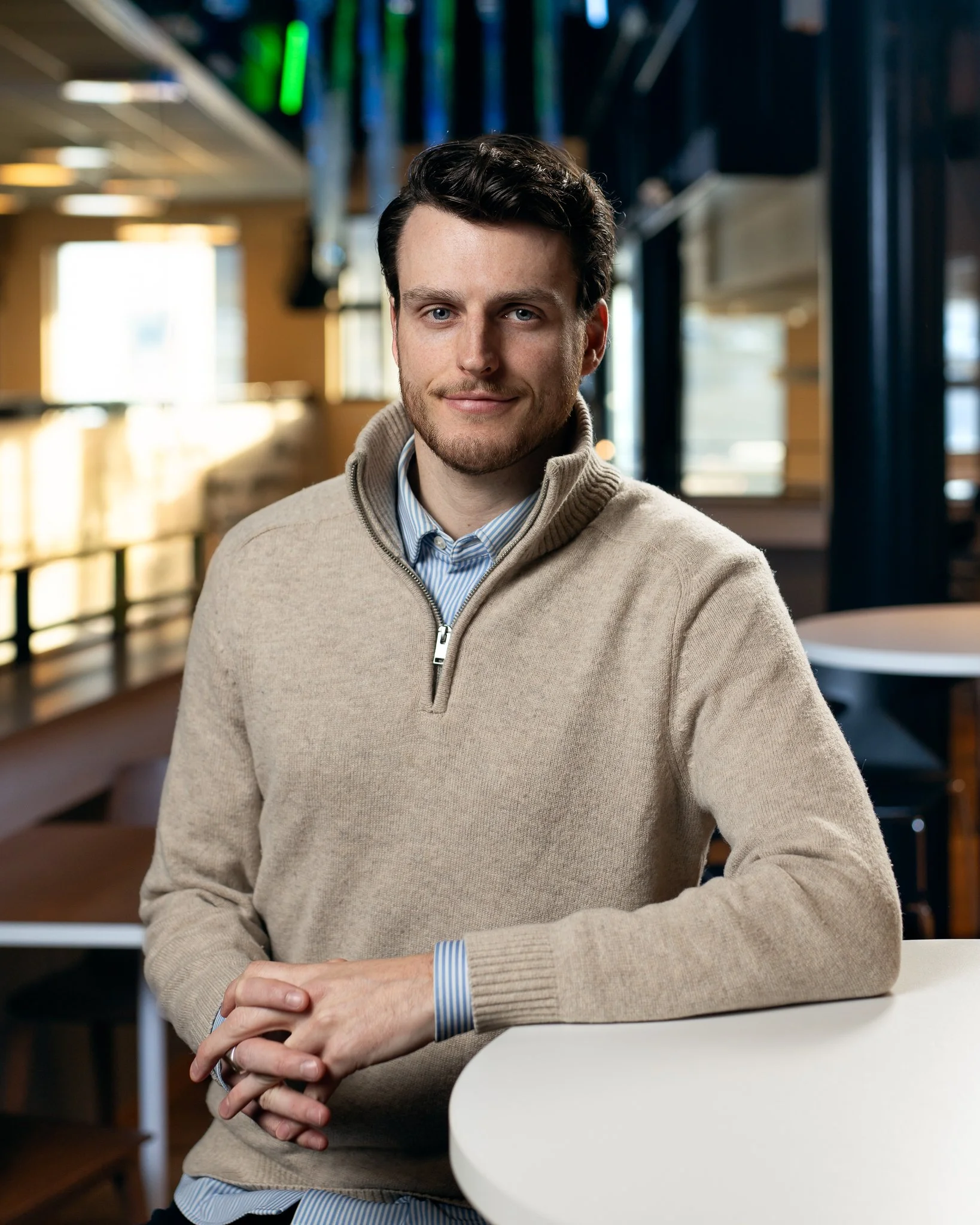 En ung mann med mørkt hår, iført en beige genser og skjorte, sitter ved et bord i en kafé eller restaurant.