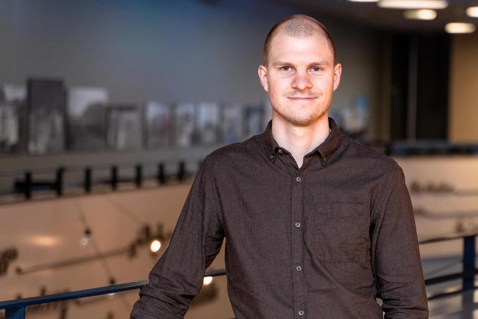 En ung mann med kort hår og skjorte står foran en bakgrunn med veggkunst eller fotografier, innendørs.