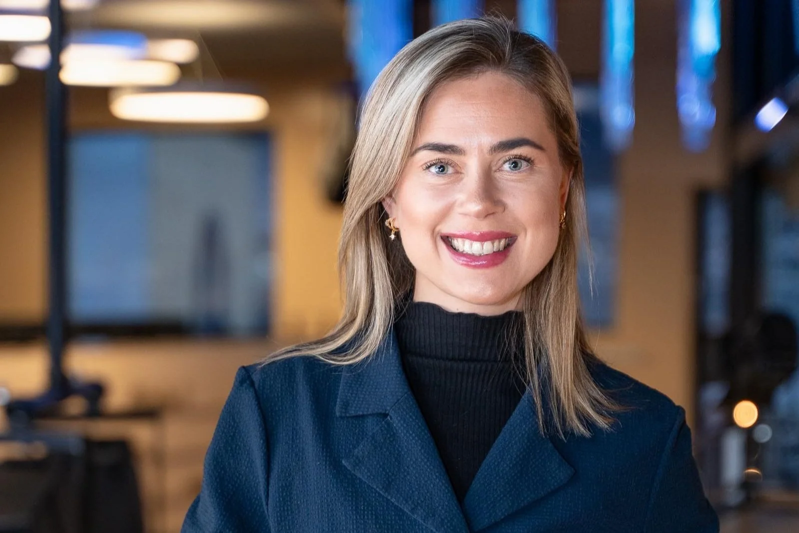 En smilende kvinne med skulderlangt blondt hår, iført mørk blazer og svart høyhalset topp, står i et moderne kontormiljø med blå og varme lys i bakgrunnen.
