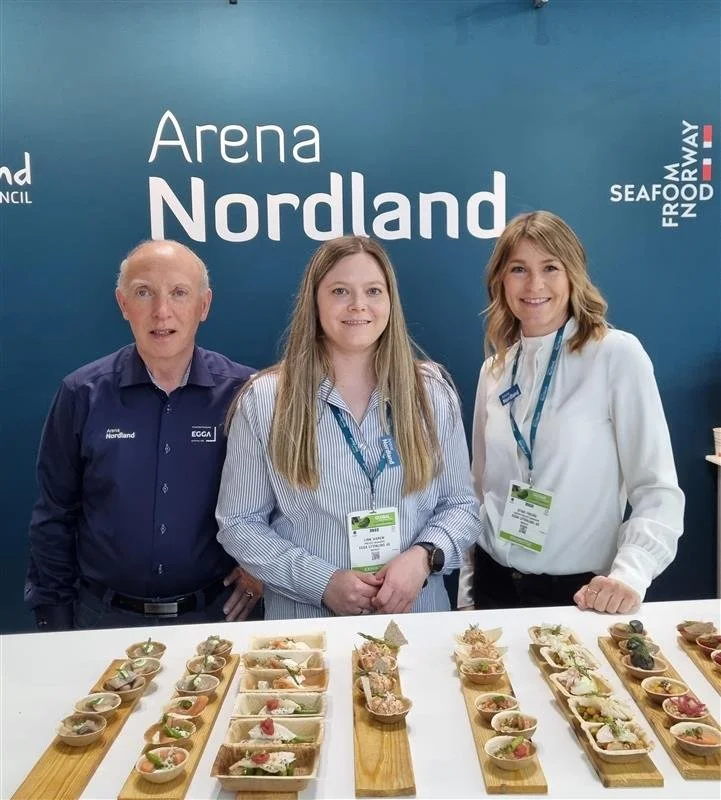 Tre personer står bak en bord med små serveringer, foran en blå vegg med teksten 'Arena Nordland' og andre logoer. Den venstre personen er en eldre mann, den midterste er en ung kvinne, og den høyre er en annen kvinne. Alle smiler, og det er tydelig