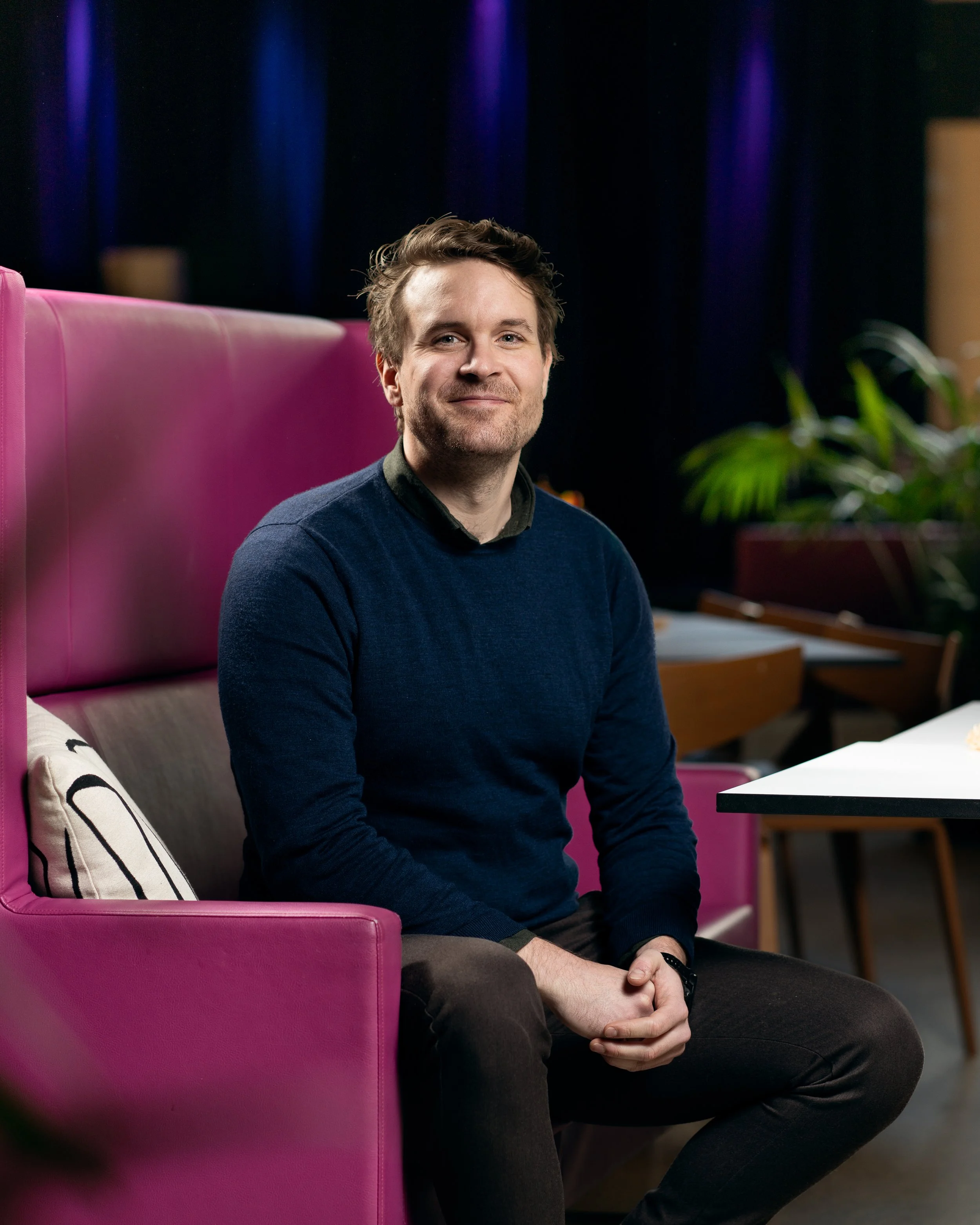 En mann sitter på en rosa lenestol i et rom med mørke gardiner og grønne planter, smilende mot kamera