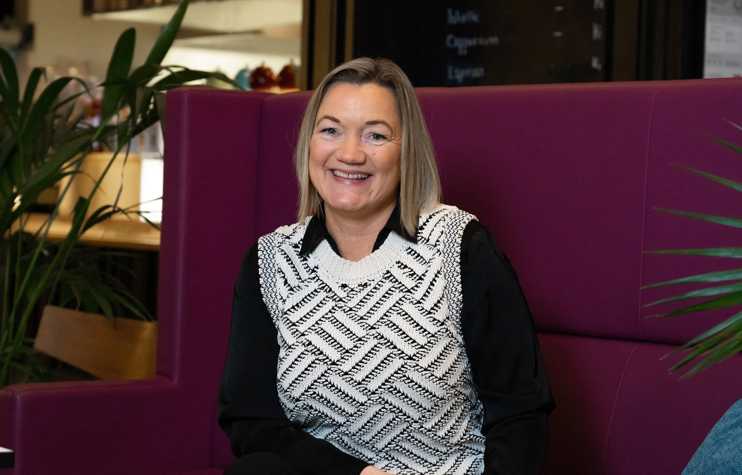 Smilende kvinne med skulderlangt hår, sitting i en kafé med rosa bøylehjørne, omgitt av grønne planter.