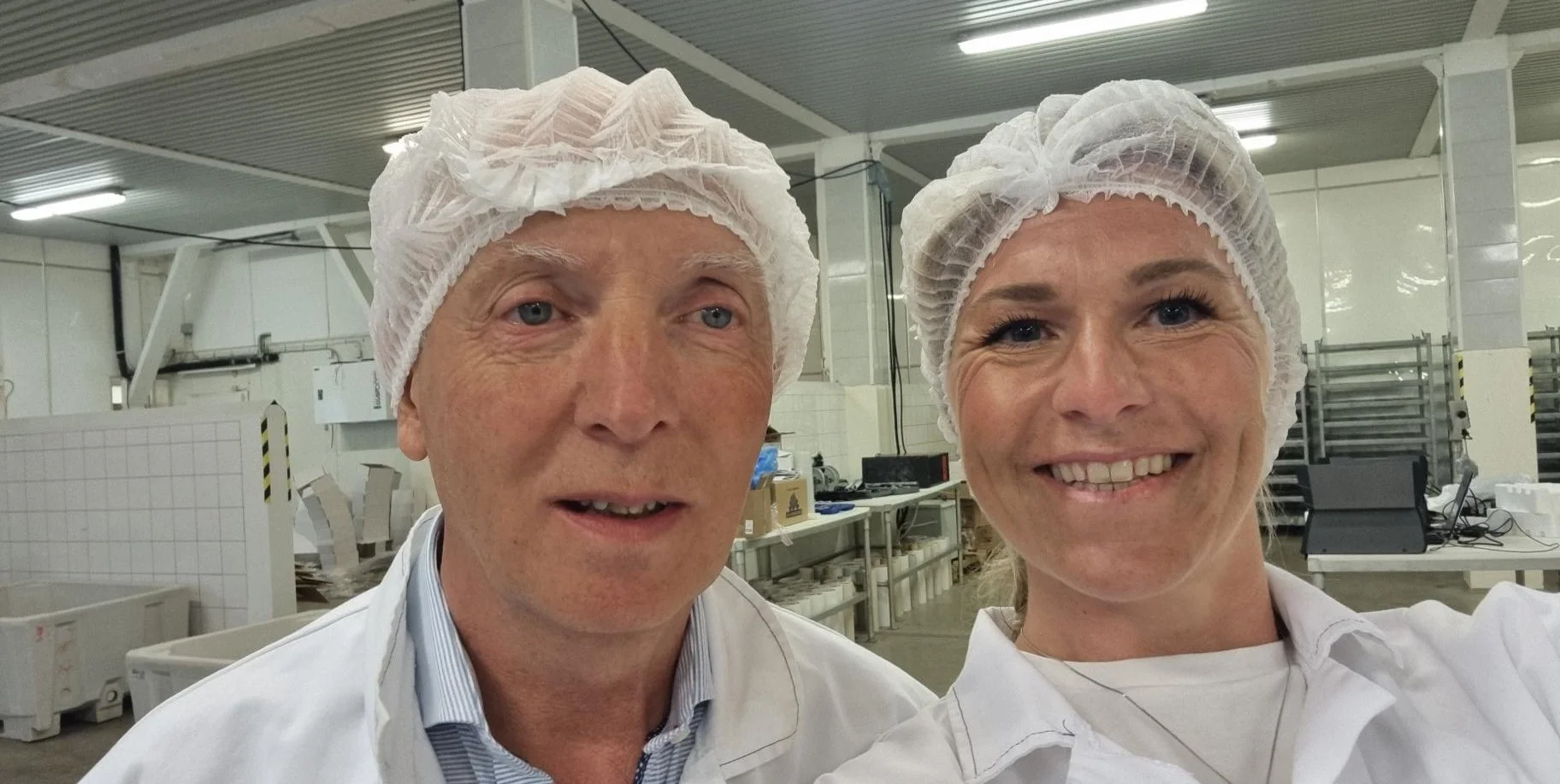 To personer iført hvite arbeidsklær og hårnetter som tar en selfie i et produksjonsmiljø med maskiner og utstyr i bakgrunnen.
