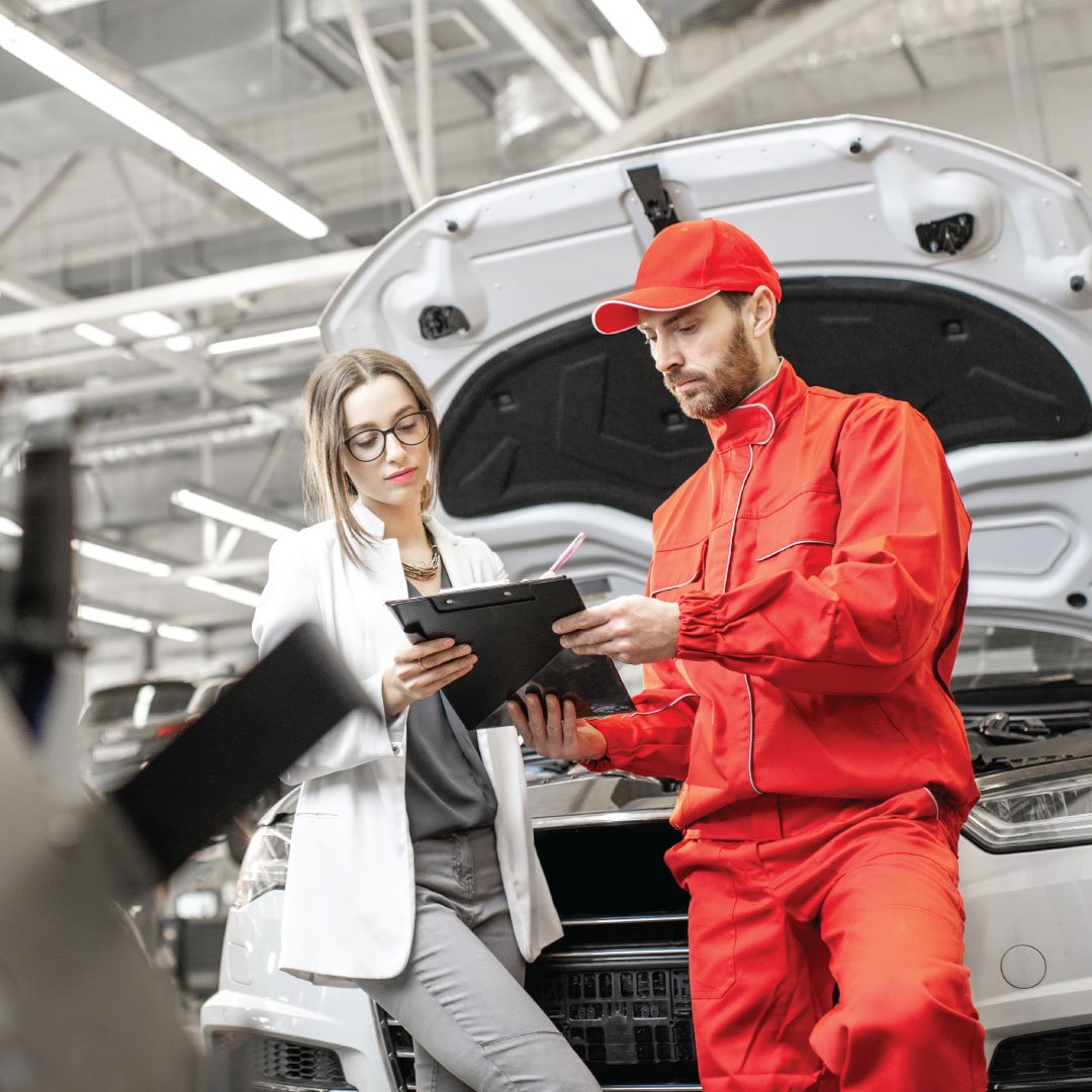 A woman and a mechanic reviewing a clipboard while standing in front of a car with the hood open inside an auto repair shop.