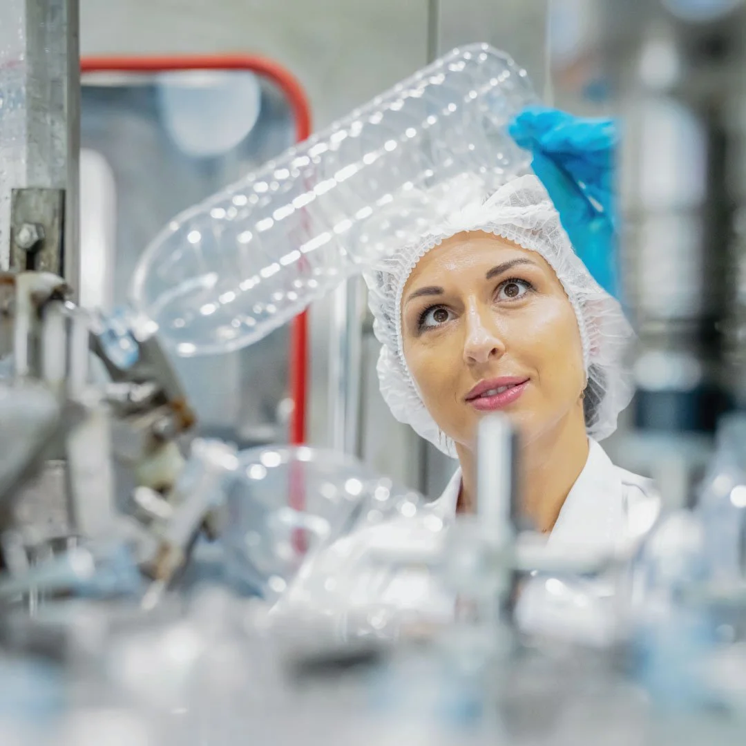 A woman in a hairnet and blue gloves working in a clean manufacturing or laboratory environment, inspecting a clear plastic bottle.