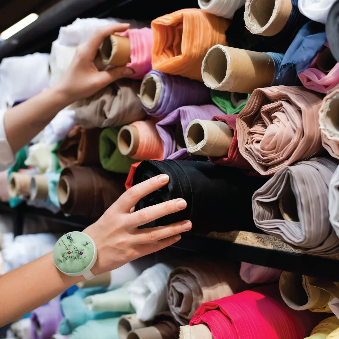 Person's hand reaching for colorful fabric rolls on a shelf in a fabric store or warehouse.