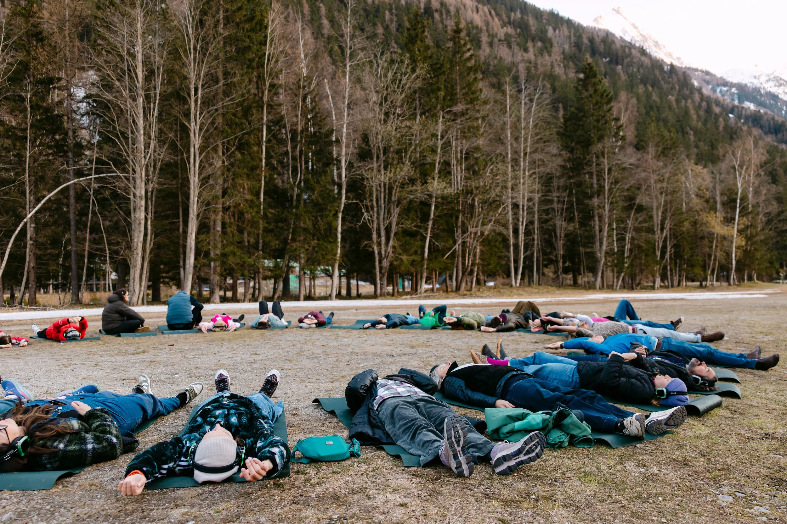 Group of people practicing yoga outdoors on mats in a field with trees and mountains in the background.