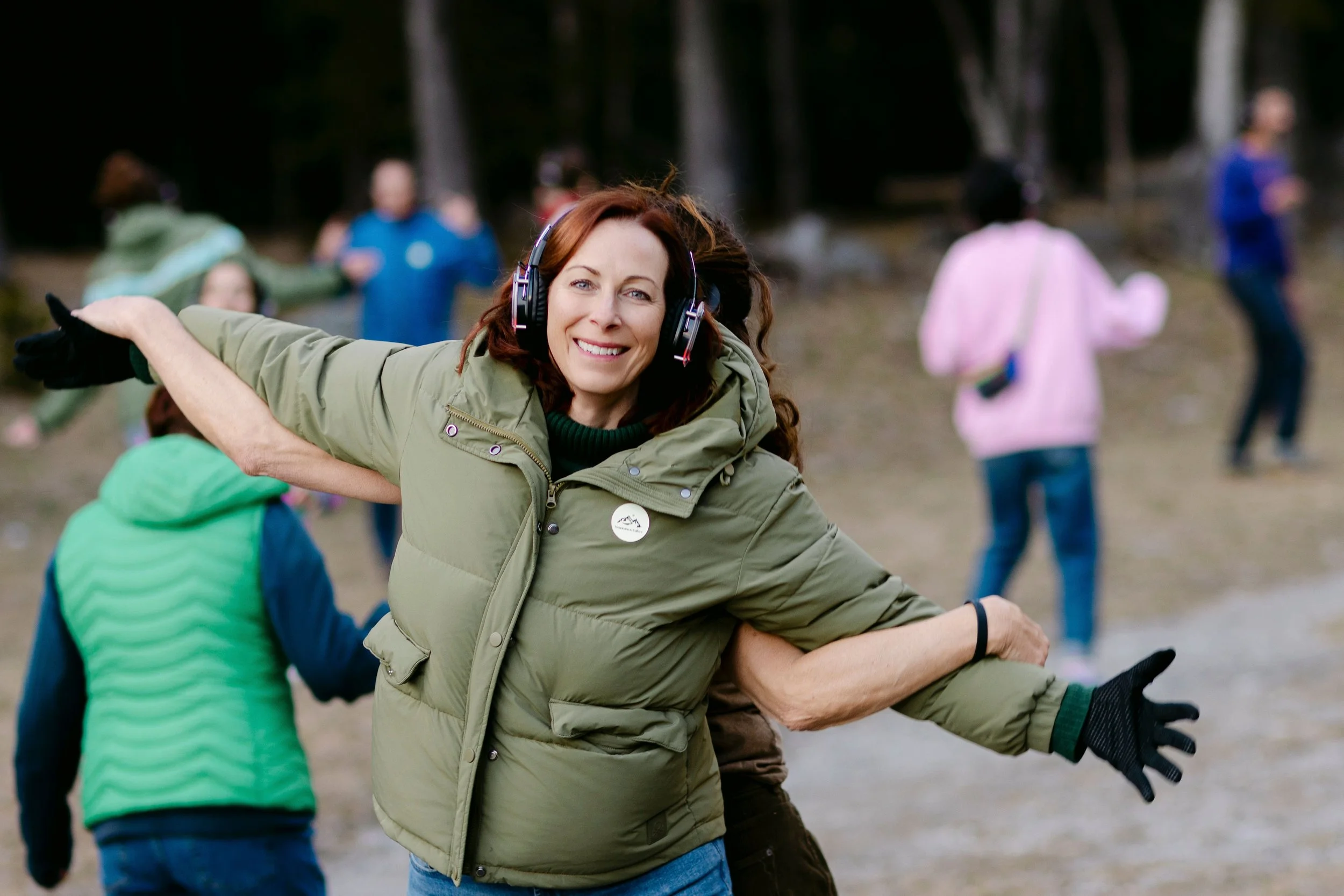 A woman with long red hair wearing headphones and a green jacket is smiling and being hugged by someone outside in a wooded area, with several other people in the background.