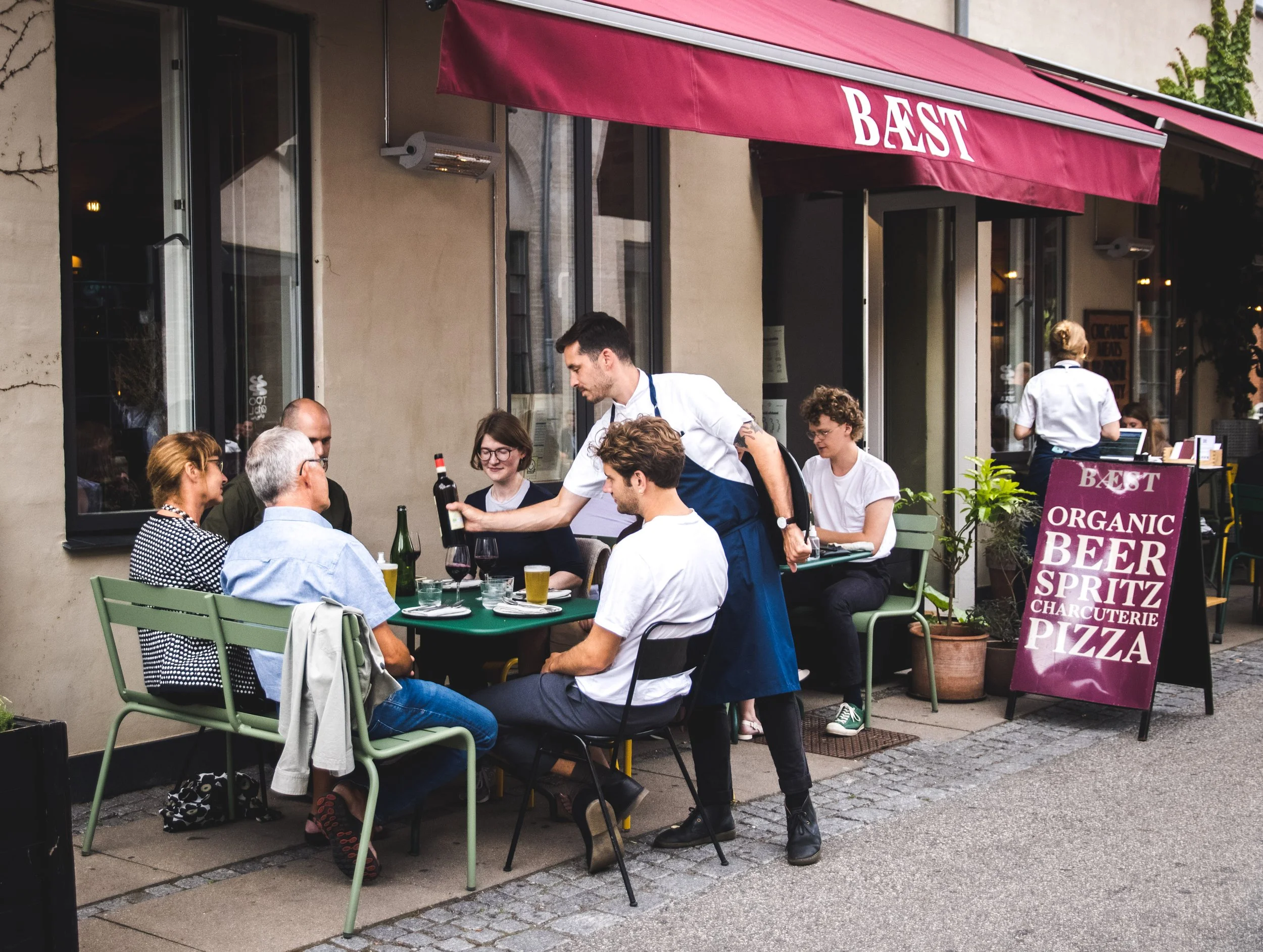 Gruppe mennesker sidder udenfor på en café ved et bord under en rød markise med teksten 'BÆST'. En tjener serverer drikkevarer. Der er en skilt med tilbud på økologiske produkter som øl, spritz, charcuterie og pizza.