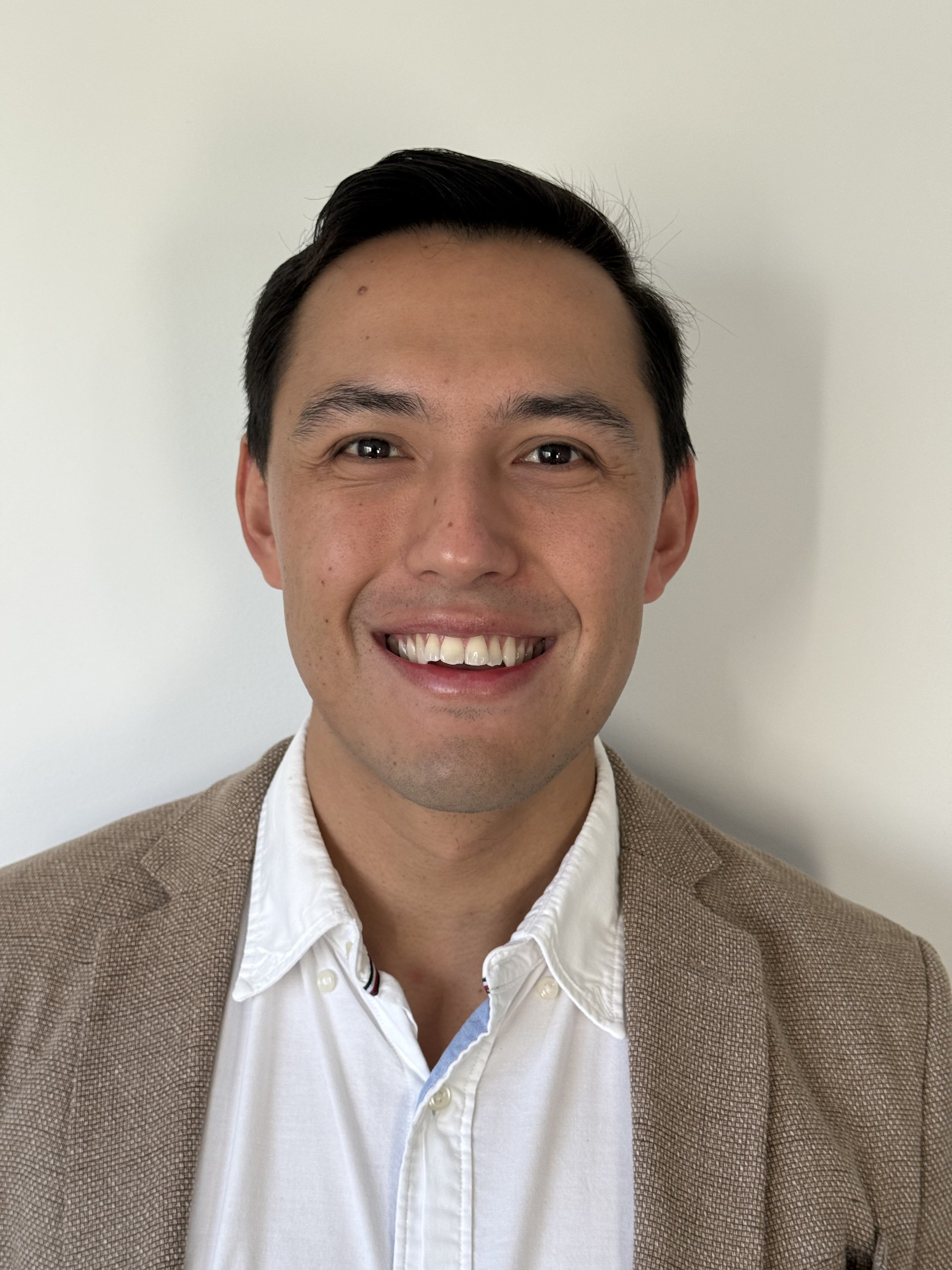 A smiling man with dark hair, wearing a white shirt and a tan blazer, standing against a plain white background. He is the reporting Sleep & Respiratory Physician of City Central Sleep Studies.