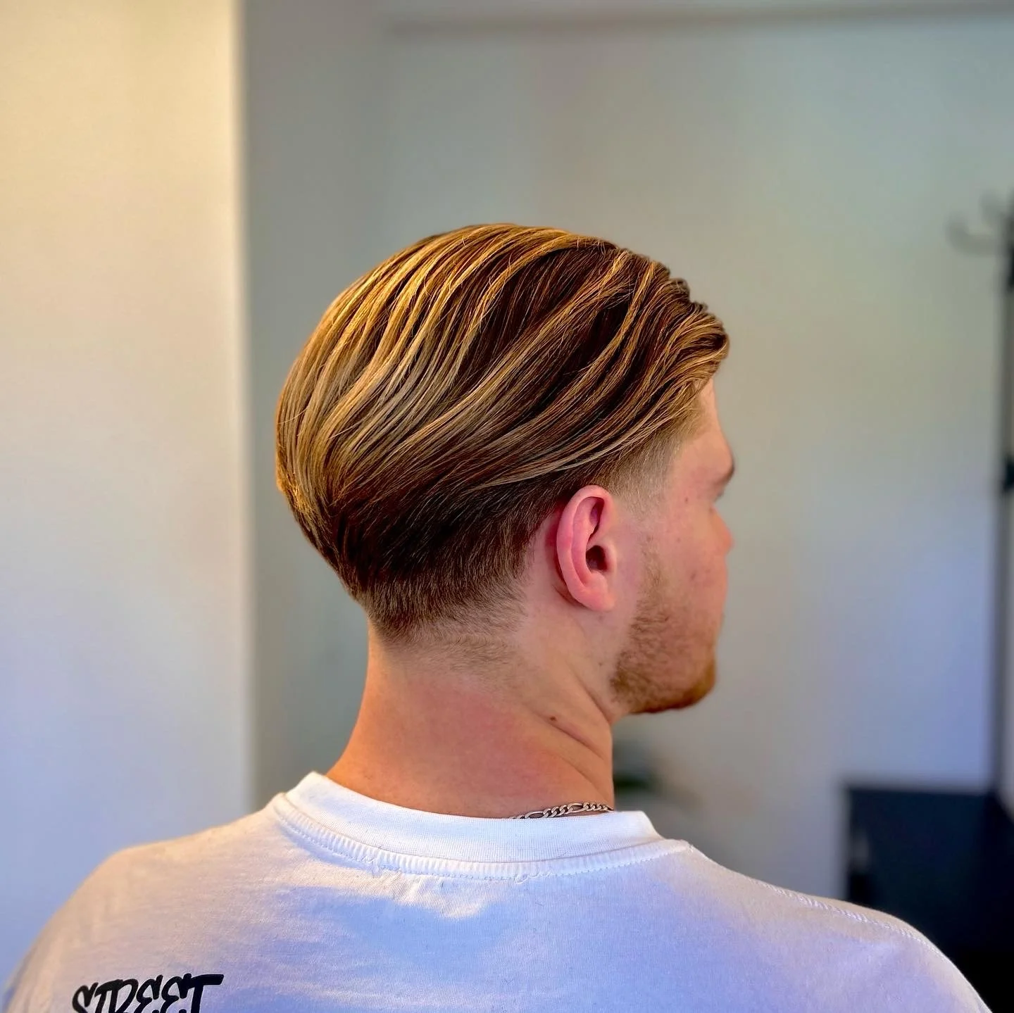 Rückansicht eines jungen Mannes mit gestyltem blondem Haar und weißem T-Shirt in einem Innenraum.