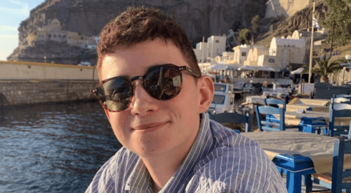 A young man with short brown hair wearing sunglasses and a striped shirt smiling outdoors near a body of water, with boats and buildings in the background.