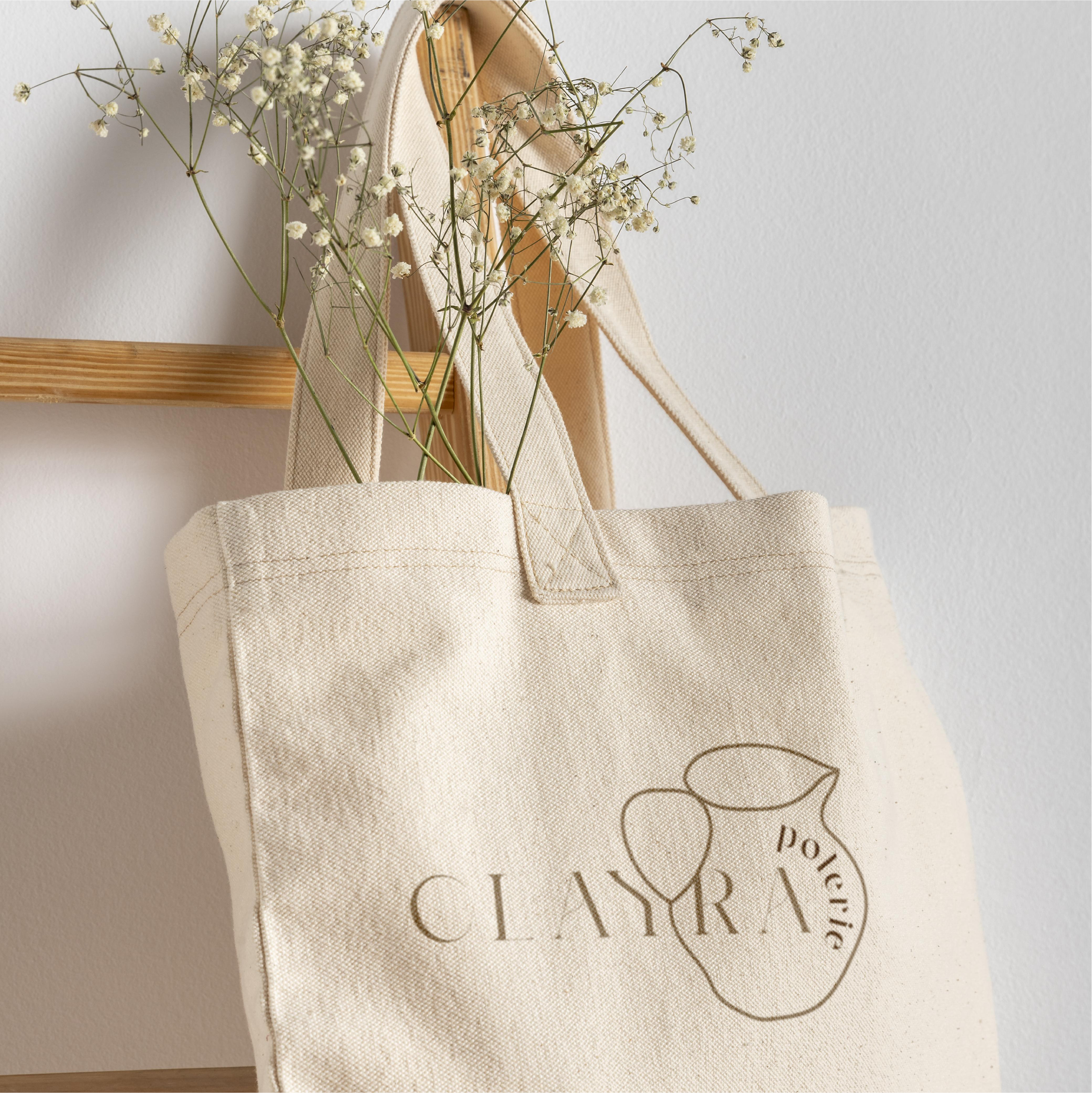 Un sac en coton beige avec un dessin de citron et le mot 'Clair' écrit dessus, suspendu à un crochet en bois, contenant des fleurs blanches légères.