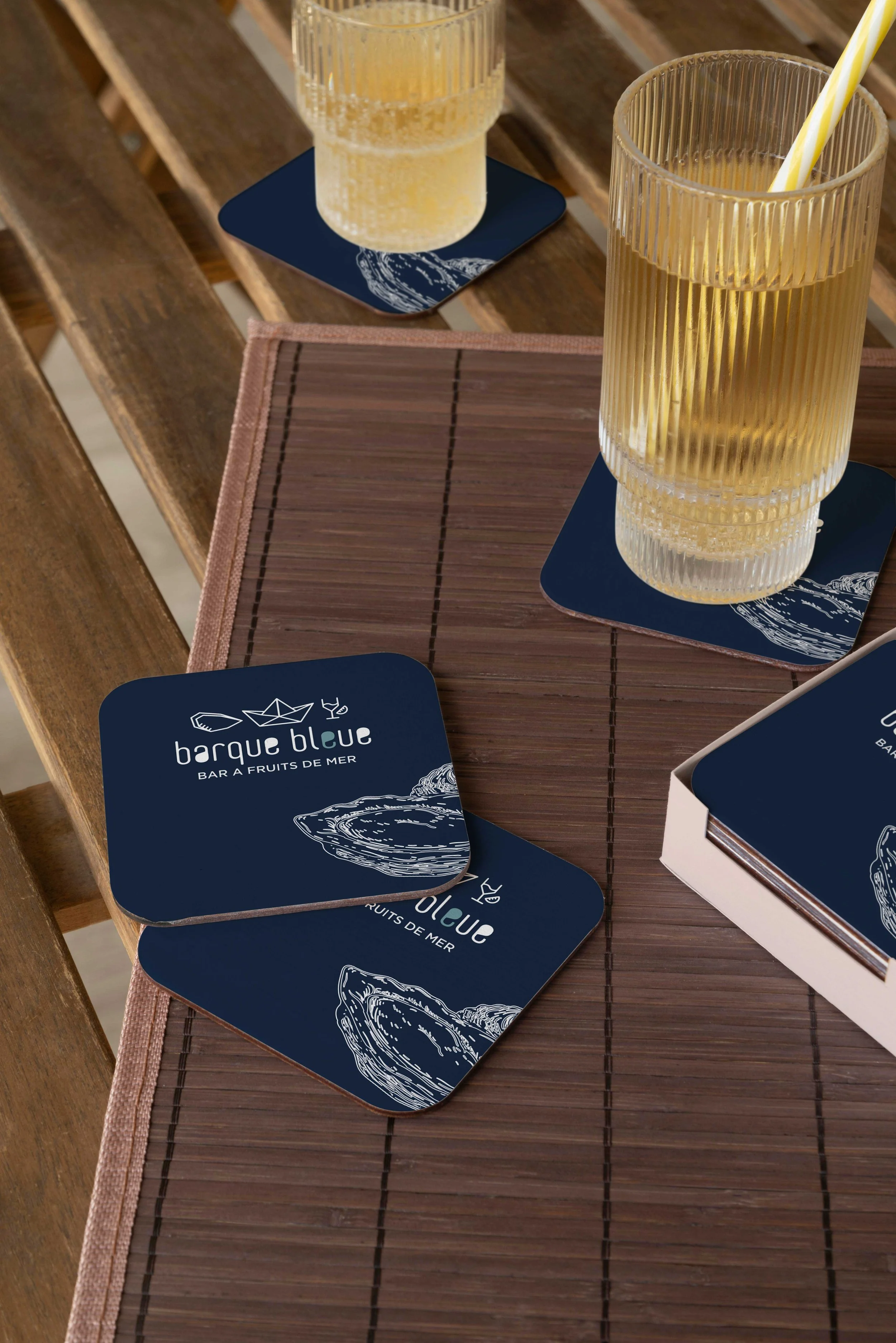 Deux verres avec une boisson jaune, chaque verre posé sur un dessous de verre bleu avec une illustration d'huître, sur une table en bois. Une boîte contenant des dessous de verre est aussi visible.