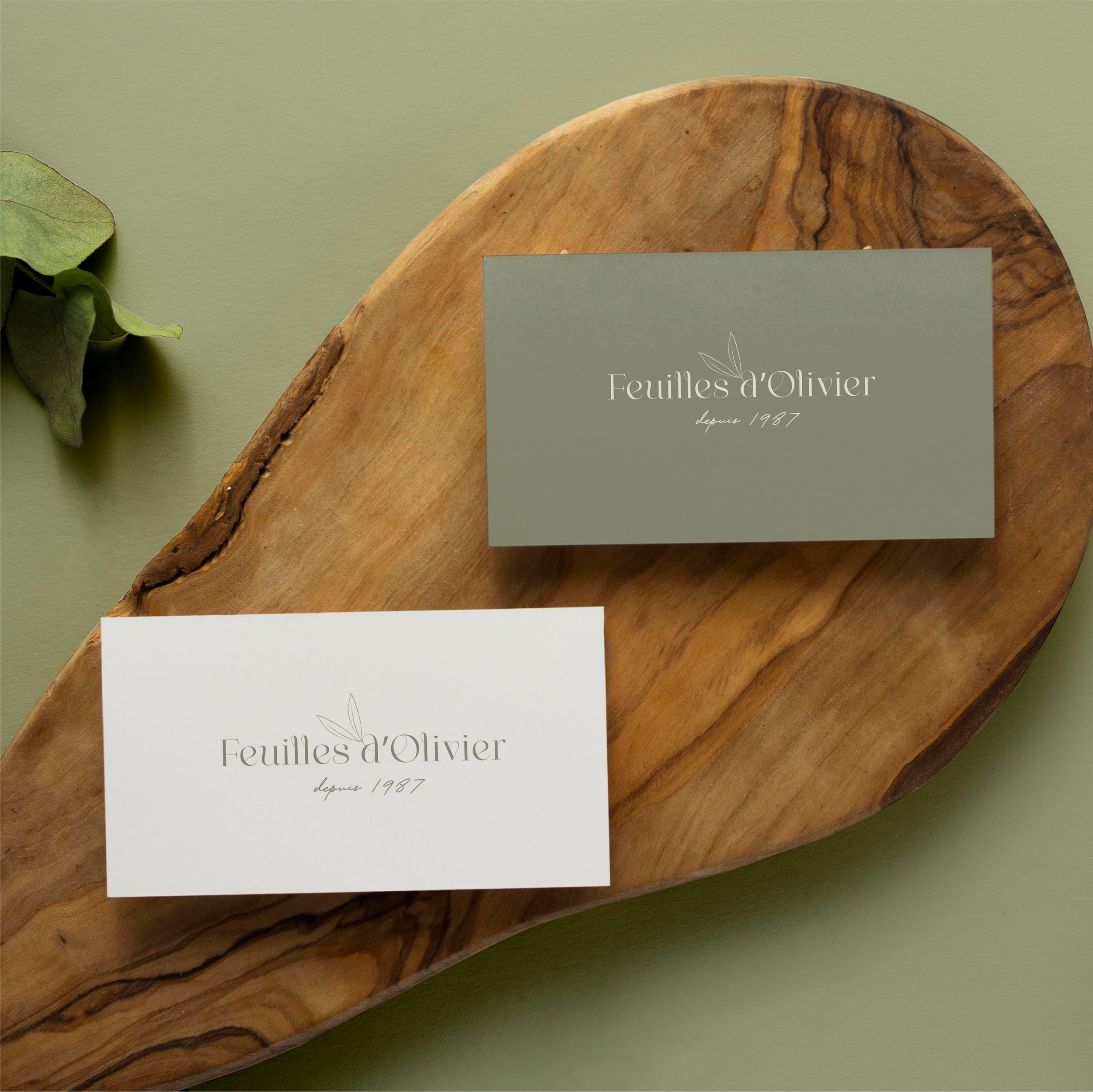 Deux cartes de visite sur une planche en bois, avec le texte "Feuilles d'Olivier depuis 1987" et une petite illustration de feuille. Une feuille verte est partiellement visible à gauche.