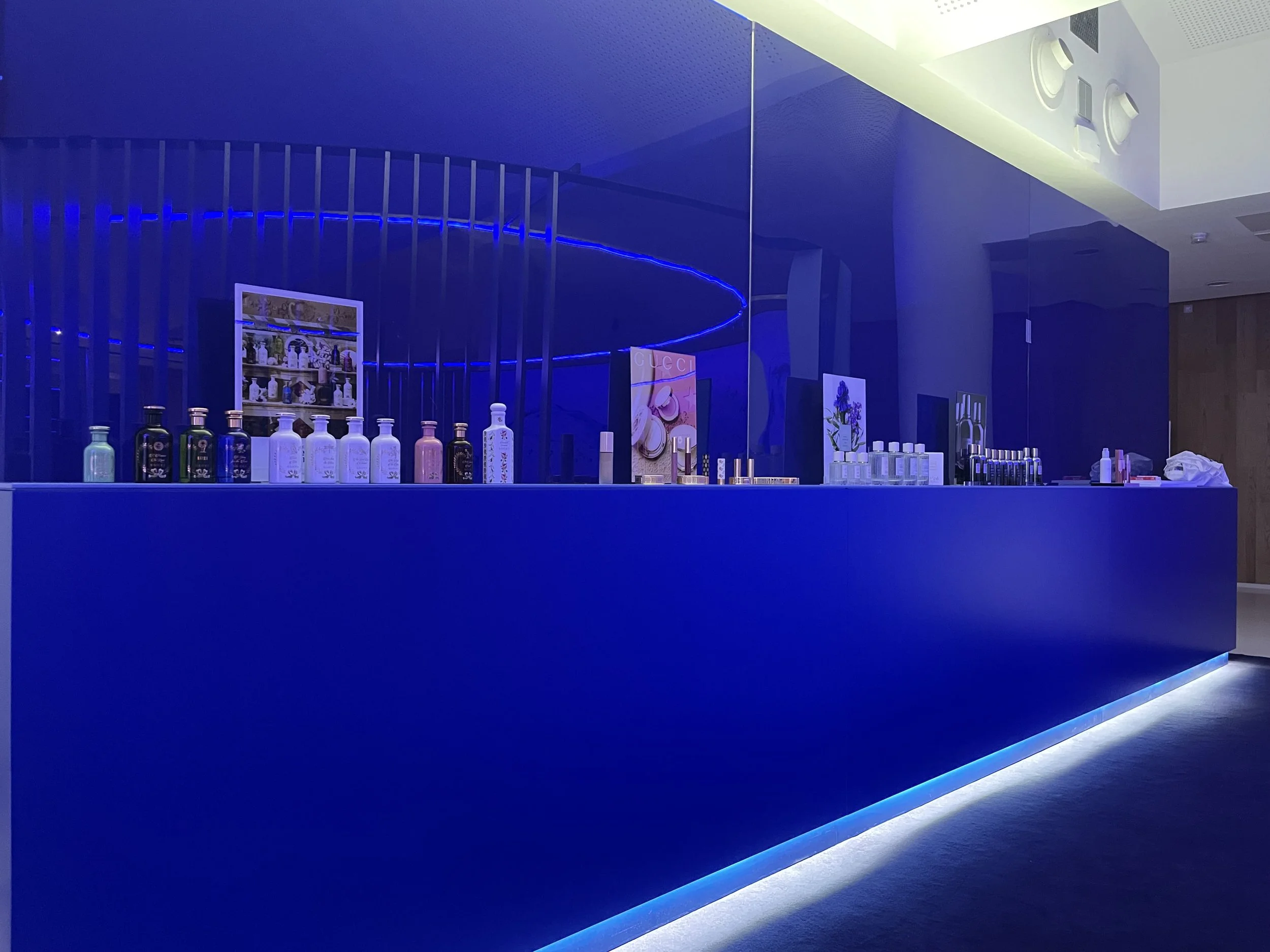 A modern bar counter illuminated with blue LED lighting, with bottles and promotional posters displayed on top and a large mirror reflecting the scene.