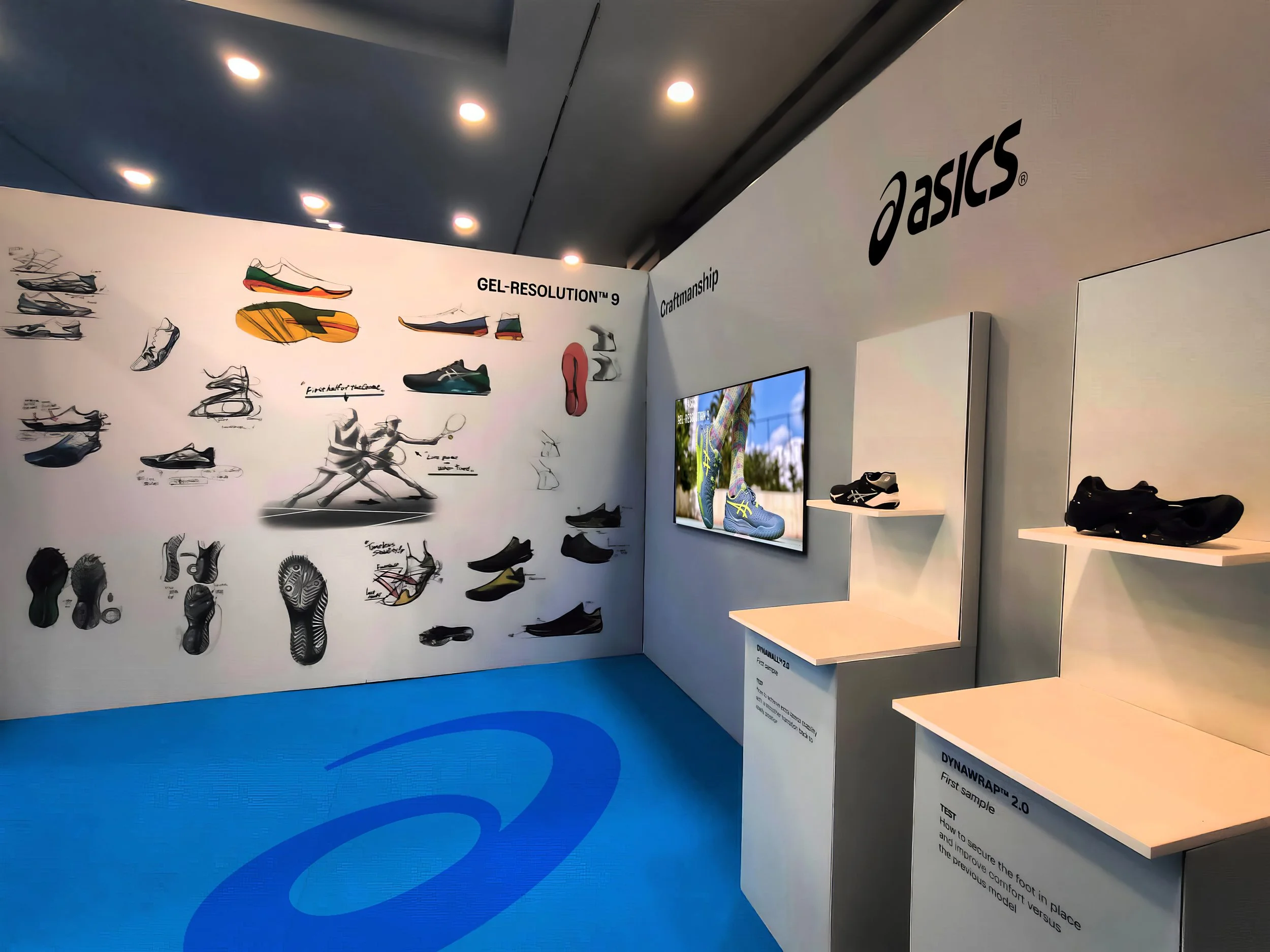 Display of ASICS running shoes with a wall showcasing shoe design sketches, a digital screen showing shoe features, and shelves with black shoes on a white background in a store in Marbella, Spain.