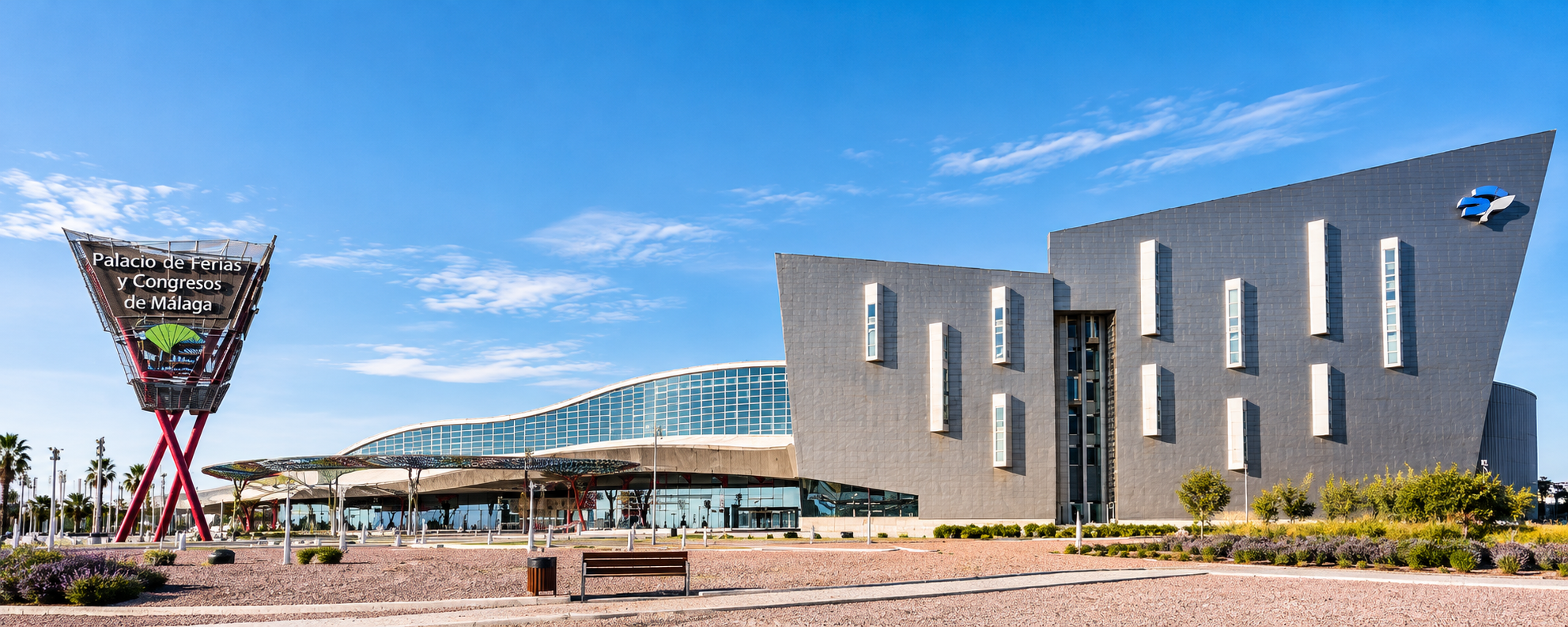 Modern conference and exhibition center with a large, angled gray facade and colorful signage that reads 'Palacio de Ferias y Congresos de Málaga.' Clear blue sky with some scattered clouds and small landscaping with bushes and trees in front.