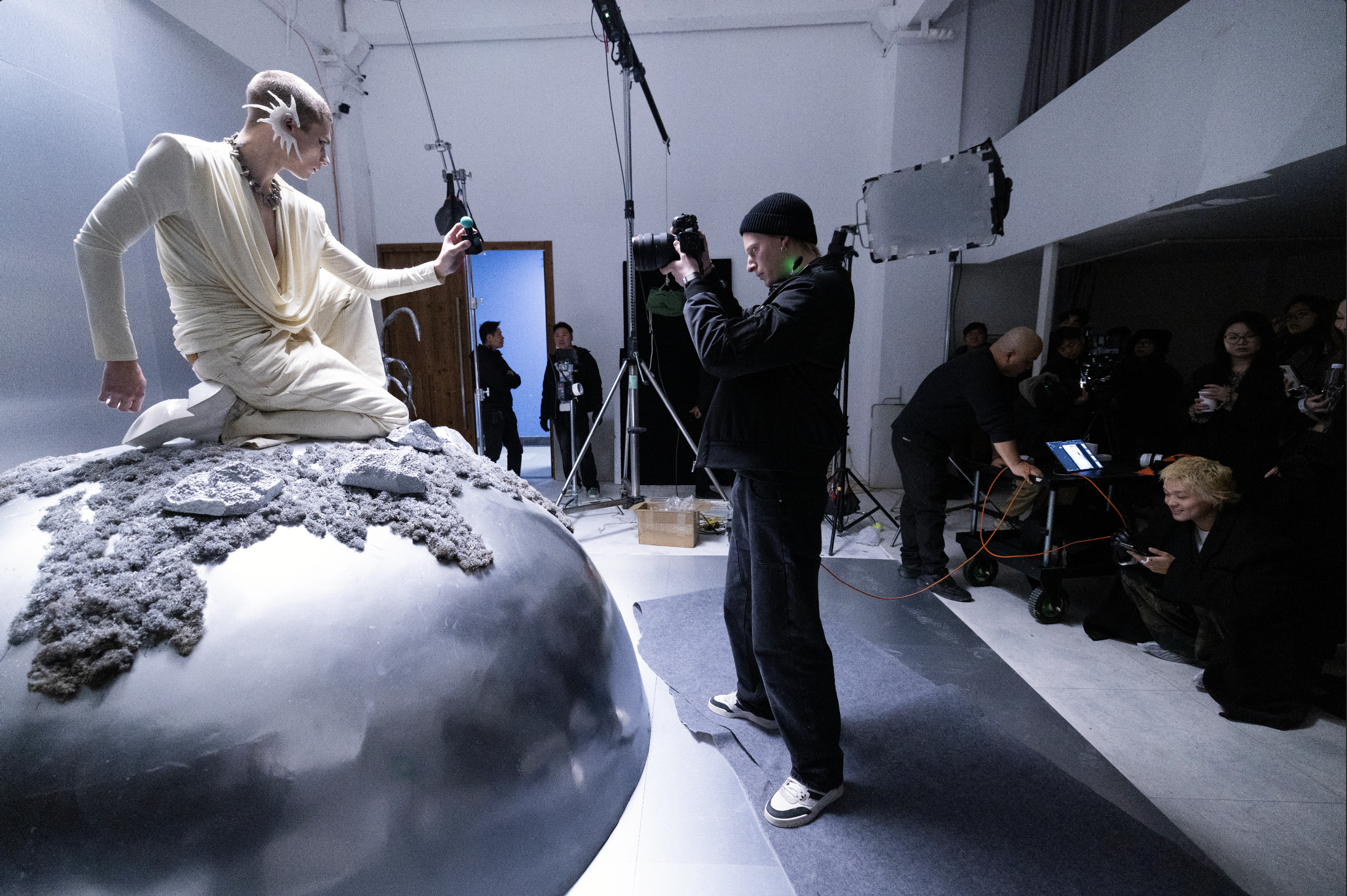 Behind the scenes of a photoshoot with a model kneeling on a large reflective sphere, wearing a cream-colored outfit with unique earrings. Photographers and crew members are setting up equipment around her in a studio.
