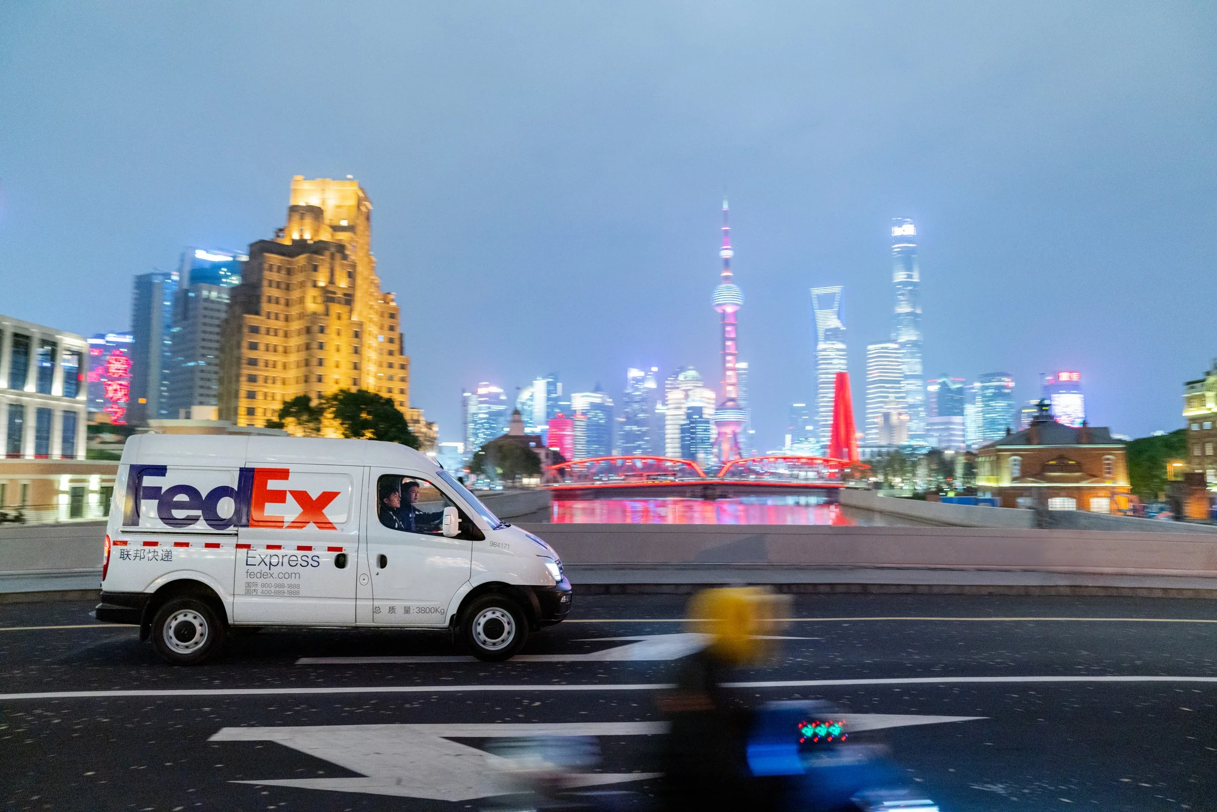 A FedEx delivery van drives on a city street at night with the illuminated Shanghai skyline, including the Oriental Pearl Tower, in the background.