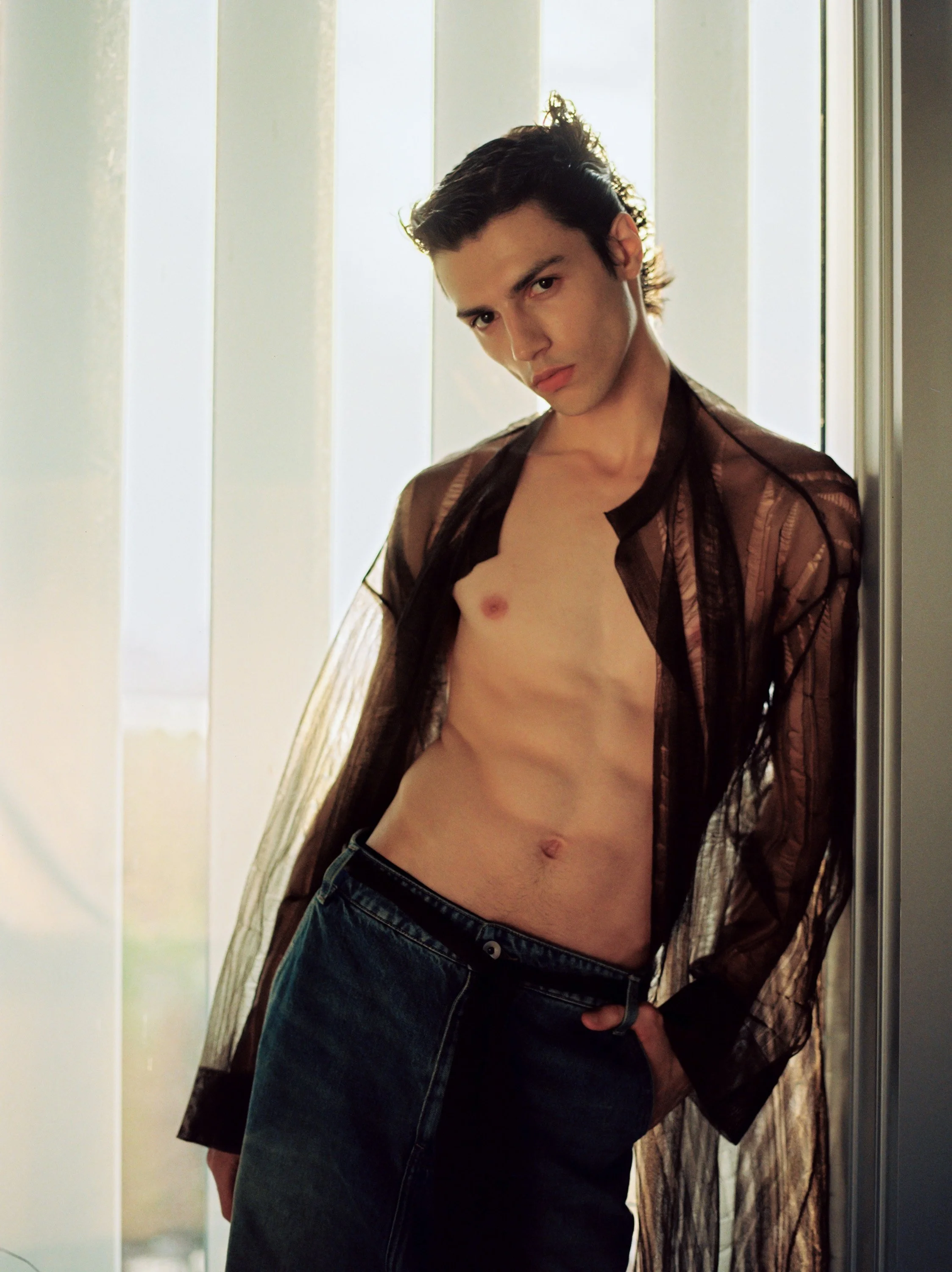 A young man with dark hair leans against a wall, wearing a sheer, brown, open shirt and dark jeans, with no shirt underneath, looking directly at the camera.