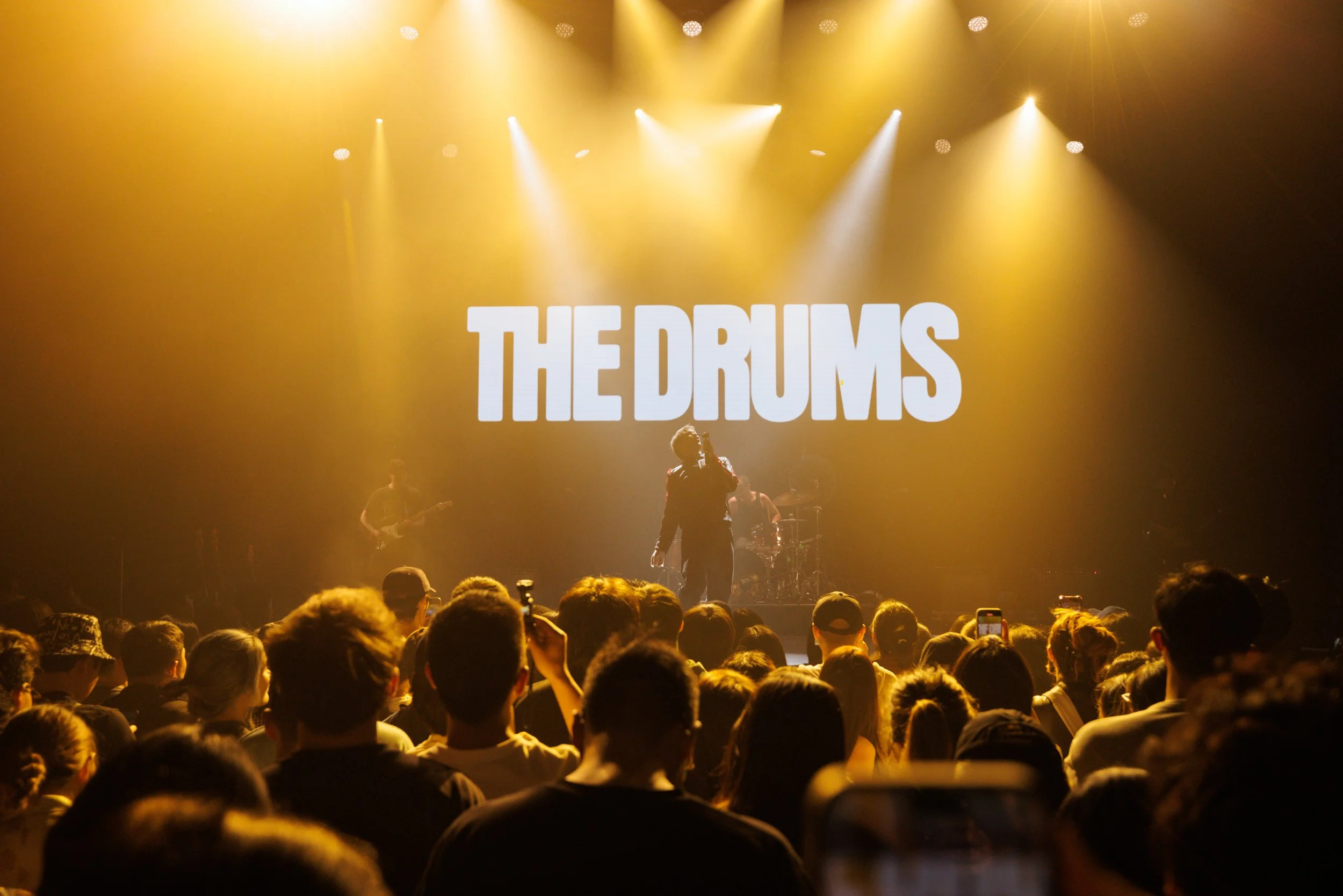 A concert scene with a band called 'The Drums' performing on stage, audience members watching and taking photos, with yellow stage lighting.