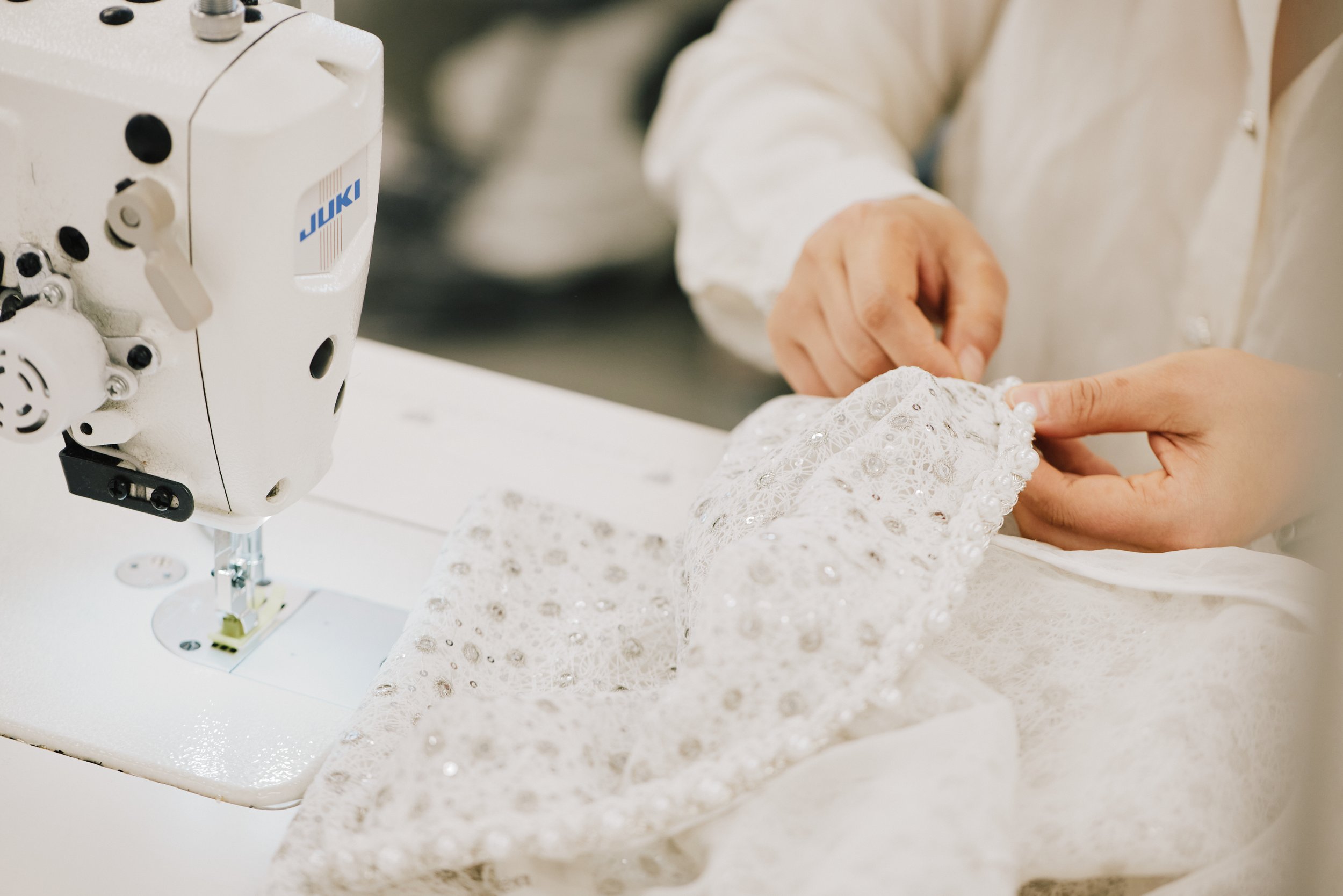 Close-up of hands guiding a sewing machine as it stitches delicate white lace fabric with intricate patterns.