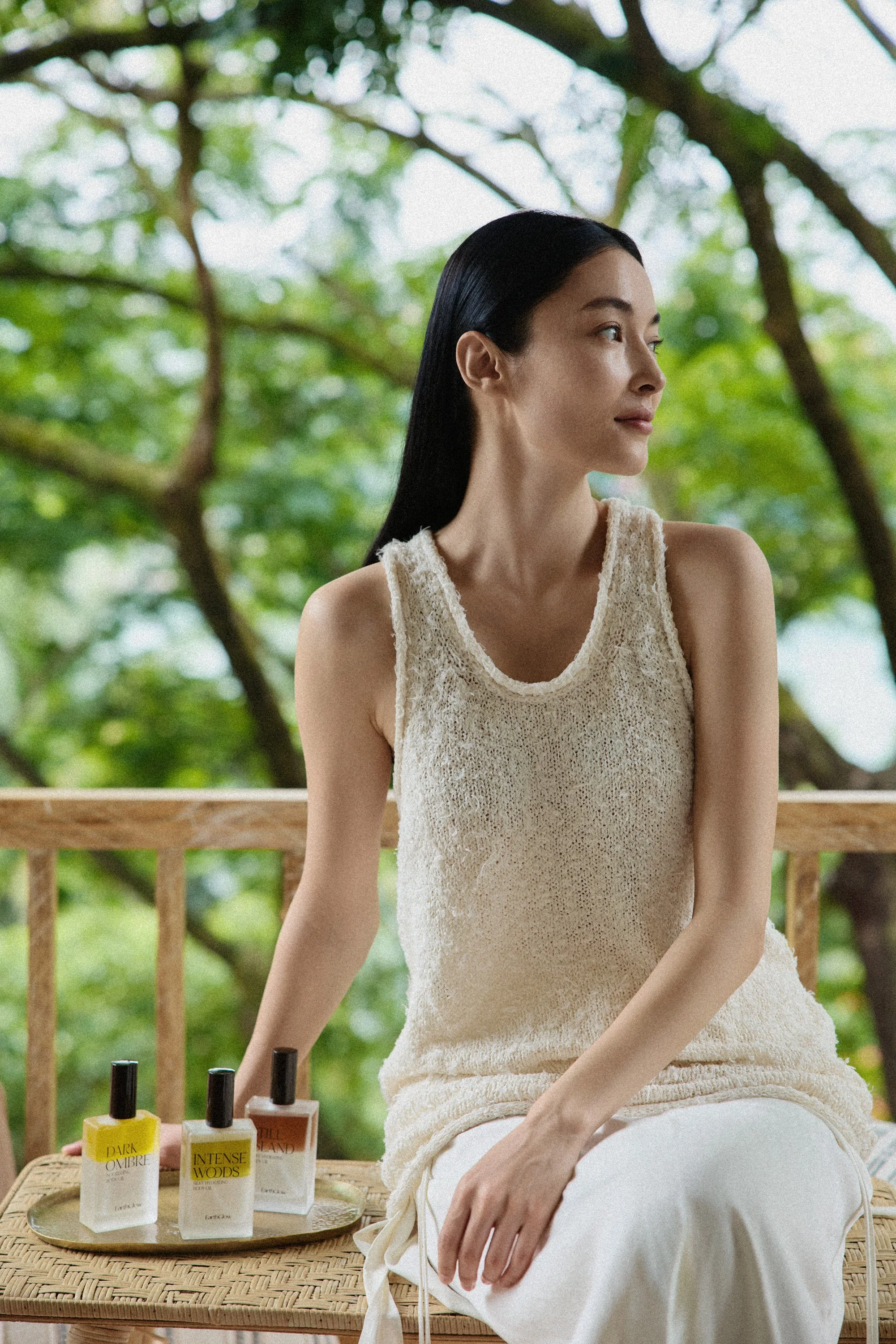A woman with long black hair wearing a beige sleeveless top, sitting outdoors on a wooden bench with trees and green foliage in the background. In front of her on a wicker table are various bottles of nail polish in yellow, beige, and brown colors.