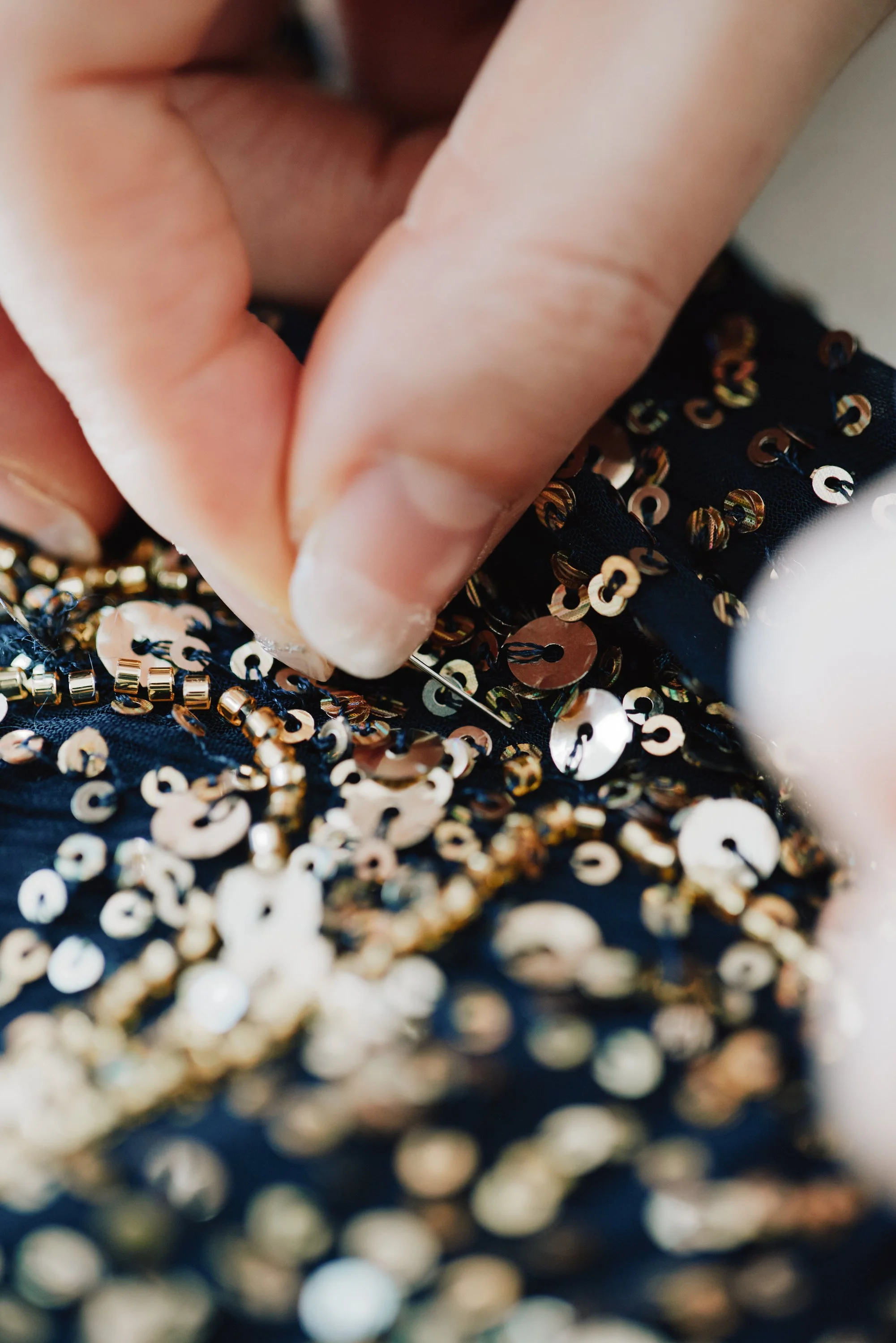Close-up of a person's hand, with fingers touching a fabric adorned with shiny, circular, gold, and pearl-colored sequins.