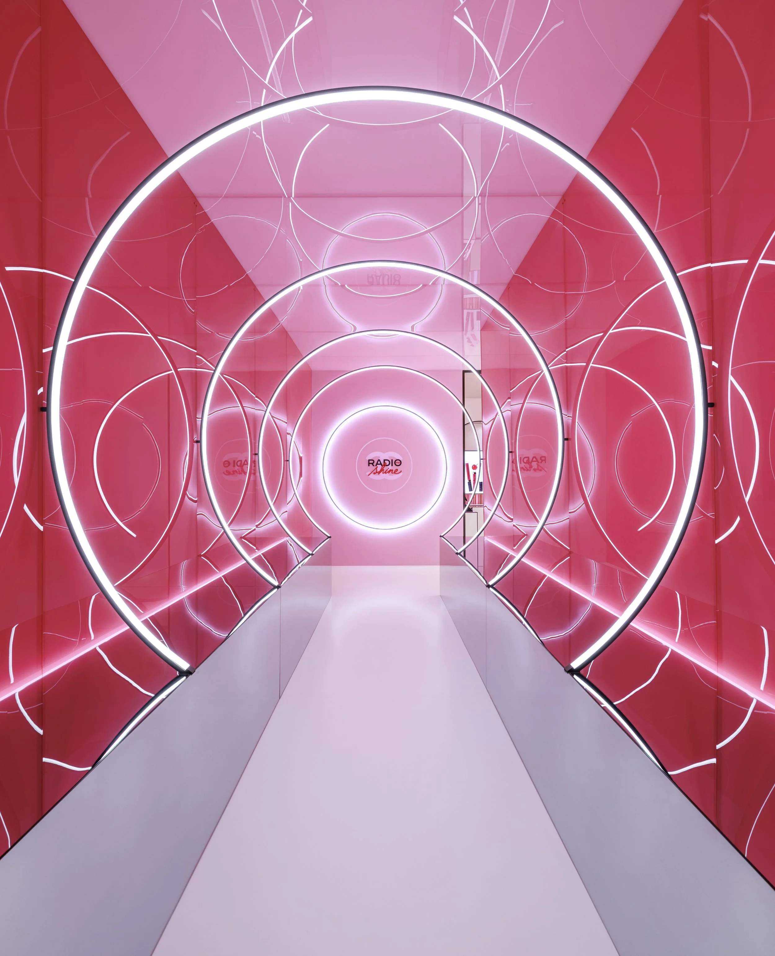 A hallway with pink walls and ceiling decorated with a pattern of illuminated white circles. Large concentric neon circles in black and white intersect, creating a tunnel-like visual effect. The floor is white, and at the end of the hallway, there is
