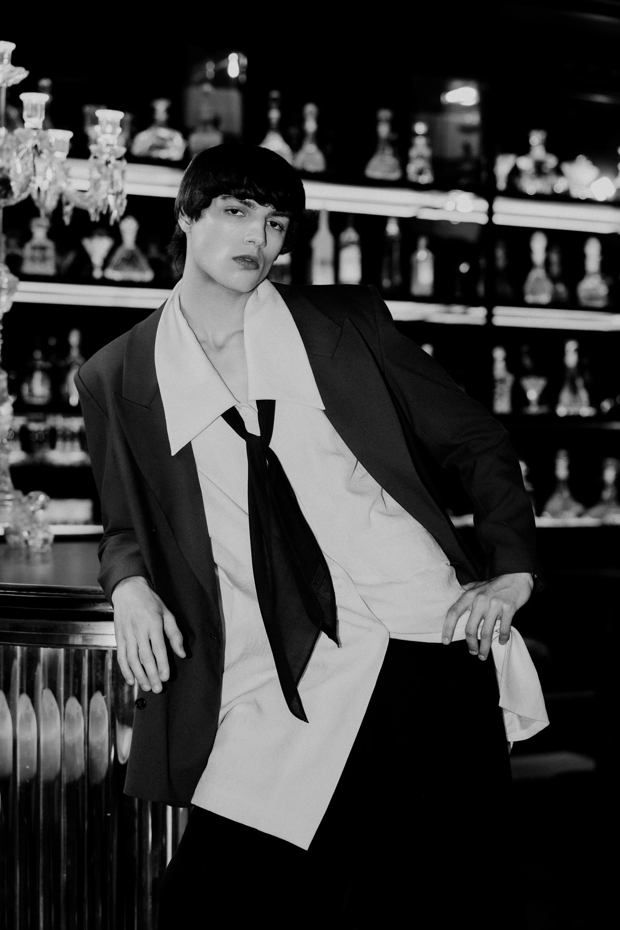 A woman with short dark hair and a serious expression, wearing a suit with a collared shirt and a tie, leaning against a bar in a dimly lit setting with bottles on shelves in the background.