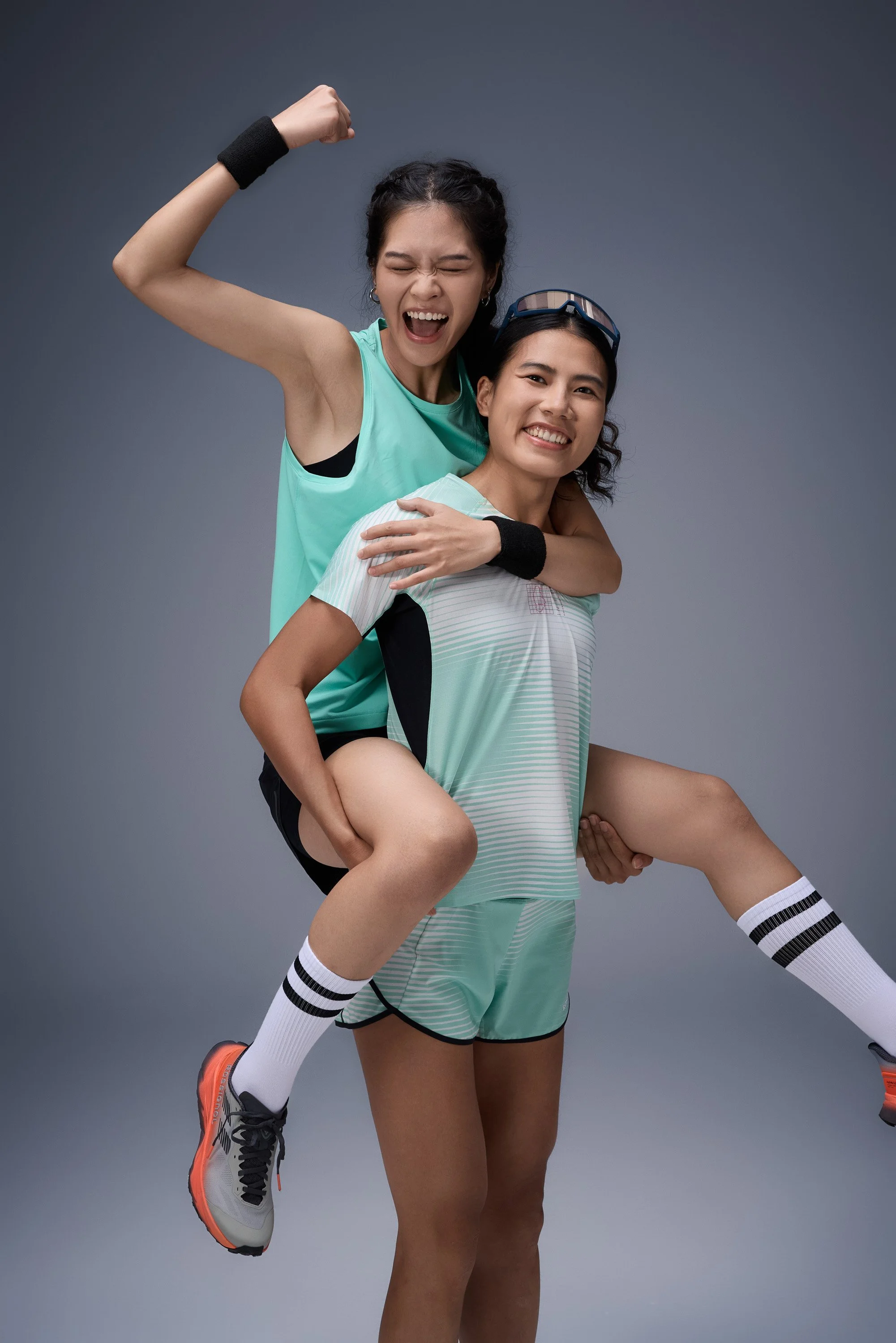 Two athletic women wearing sports clothes, one giving the other a piggyback ride. The woman in front is smiling, while the woman on the back is celebrating with her arm raised and fist clenched.