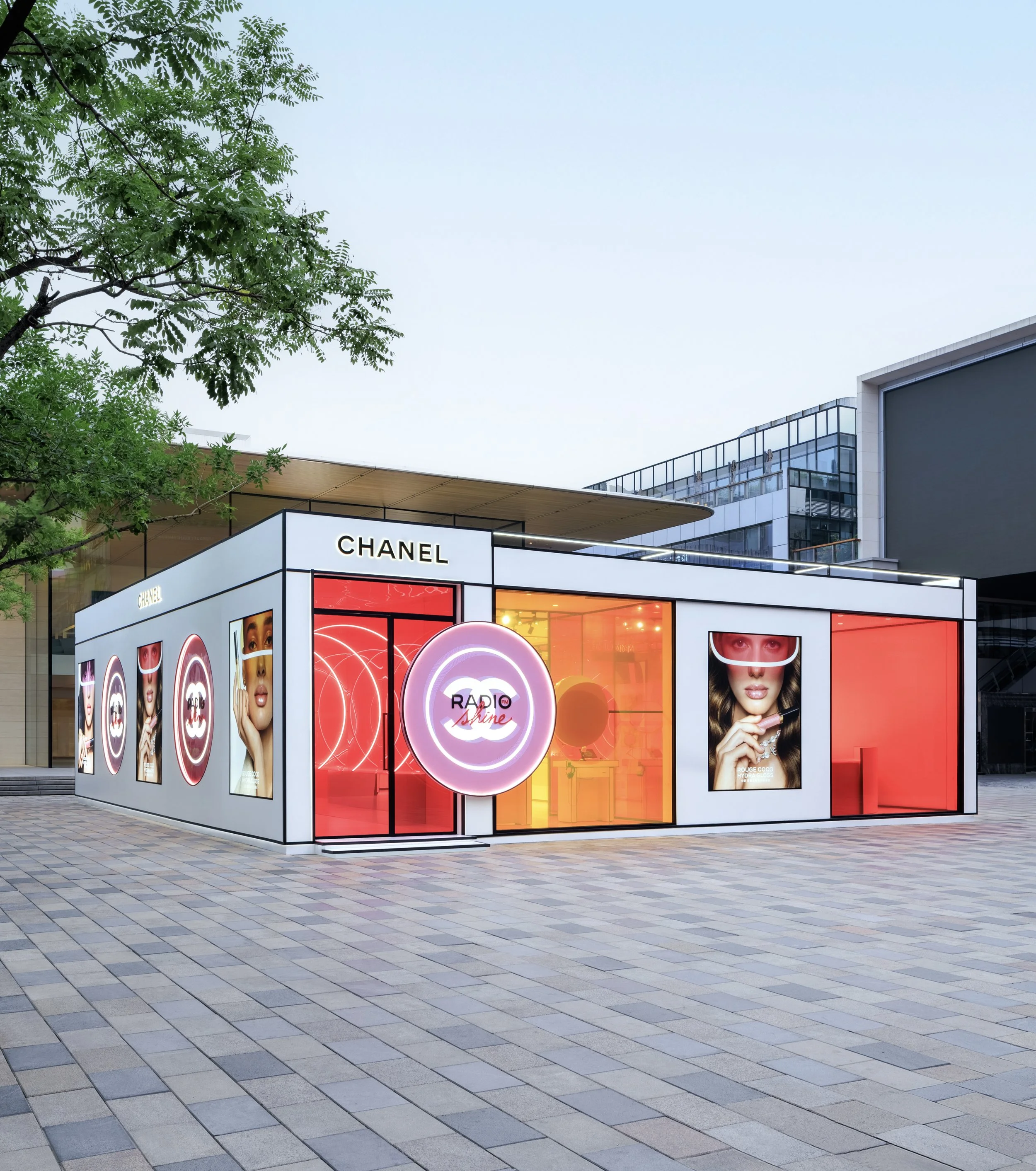 An outdoor retail kiosk with a white facade displaying advertisements for cosmetics, including images of women with makeup. The kiosk has neon signs and a central circular sign with the logo for Radiance Shine. In the background, there is a modern bu