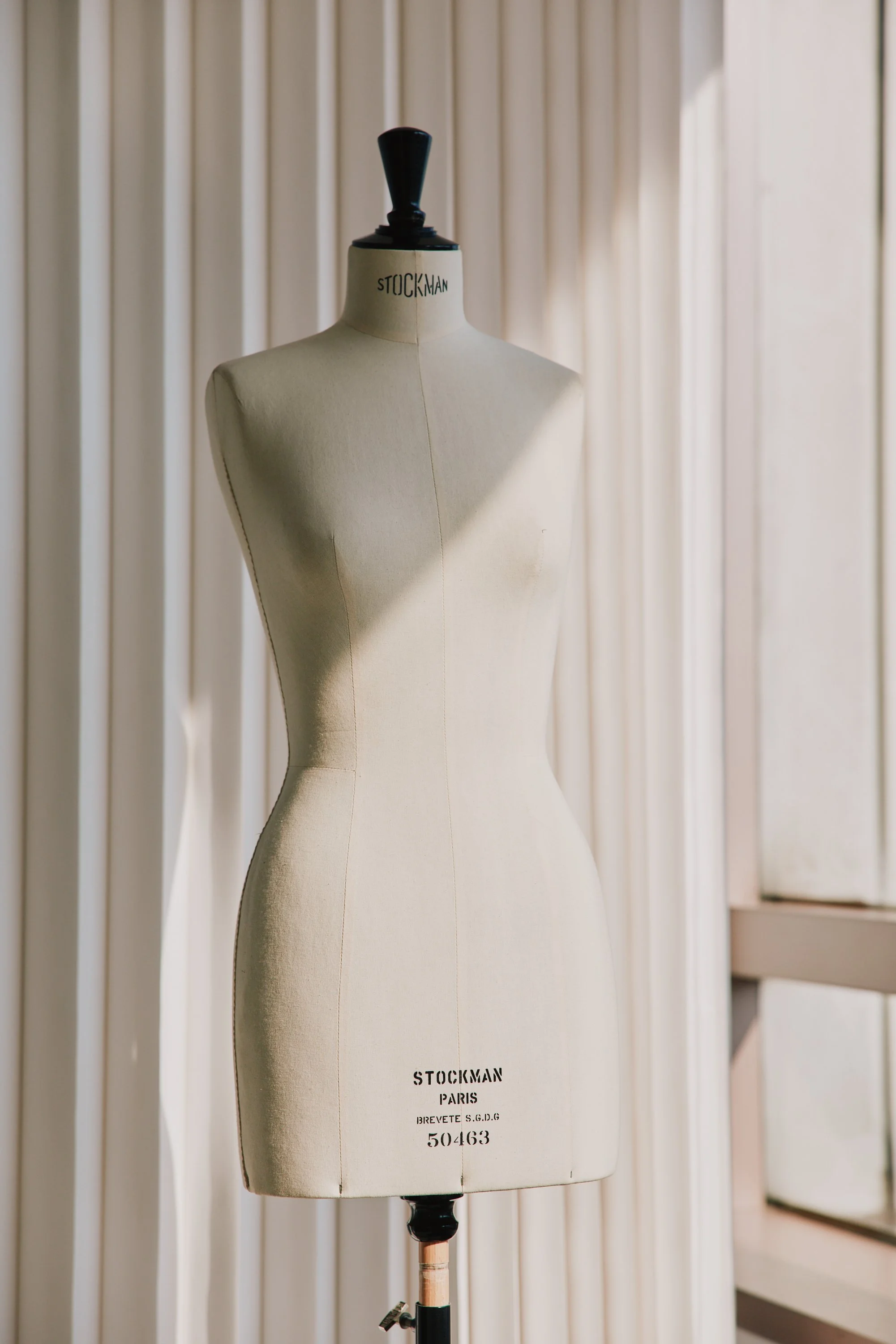 Cloth-covered dress form mannequin on a stand, labeled 'Stockman Paris,' in front of vertical blinds.