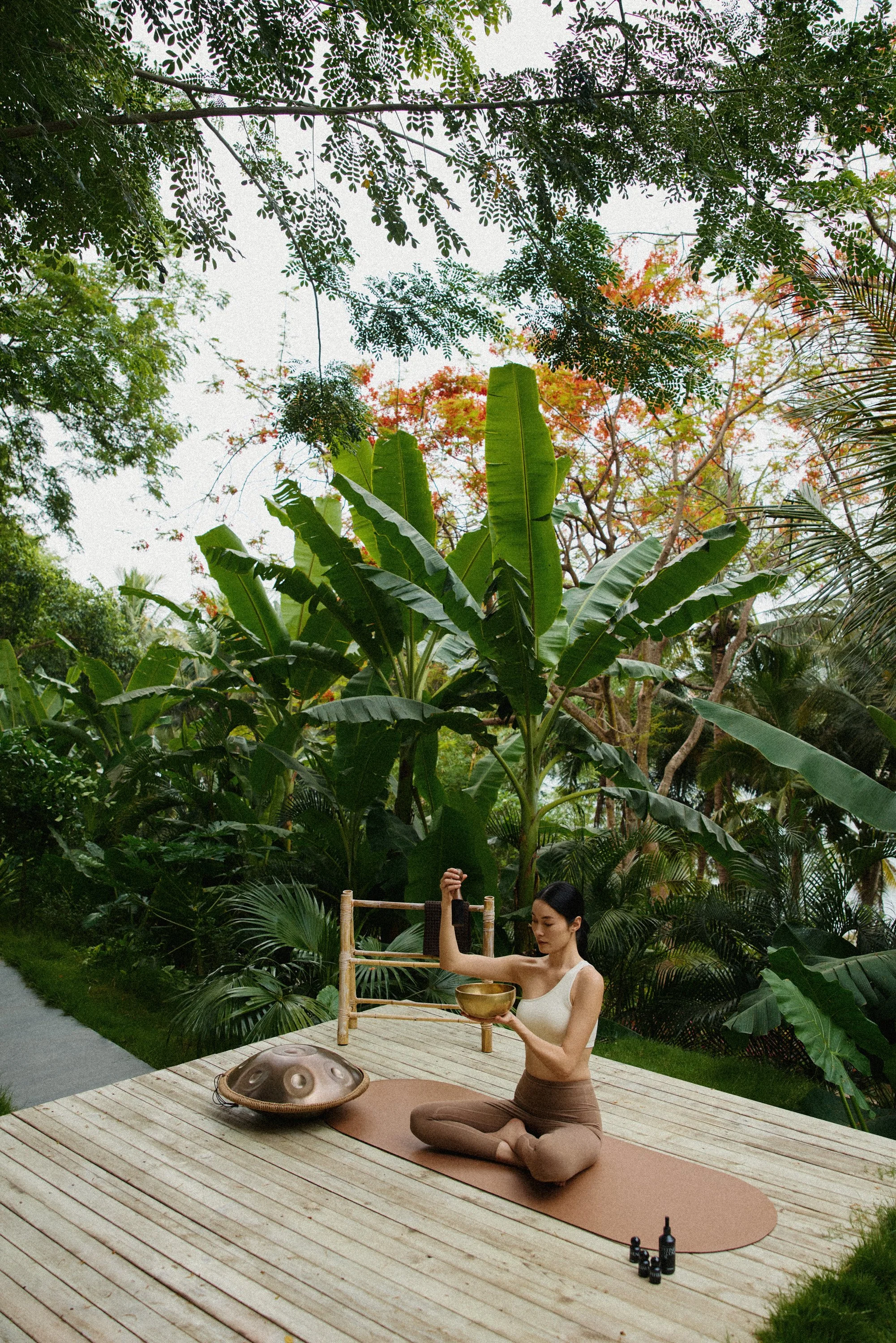 A woman practicing yoga outdoors on a wooden deck surrounded by lush green tropical plants and trees, holding a singing bowl in one hand with her eyes closed.