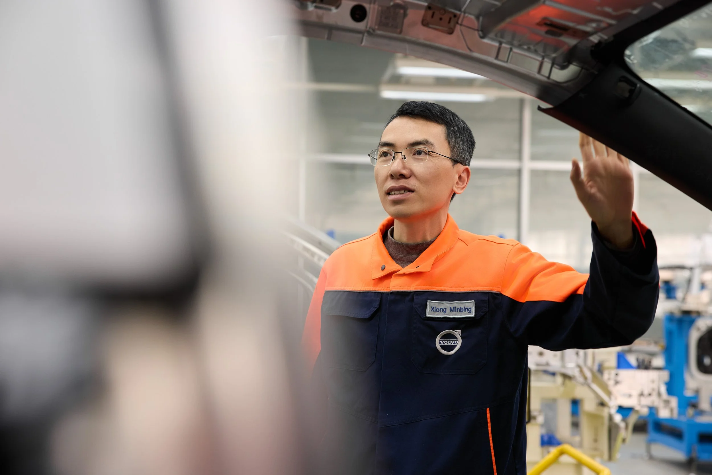 A man wearing glasses and an orange and navy jacket with a Volvo logo and a name tag reading 'Xiong Minbing' is working in an industrial or manufacturing setting. He is looking up and slightly to his left with his right hand raised, possibly inspecti