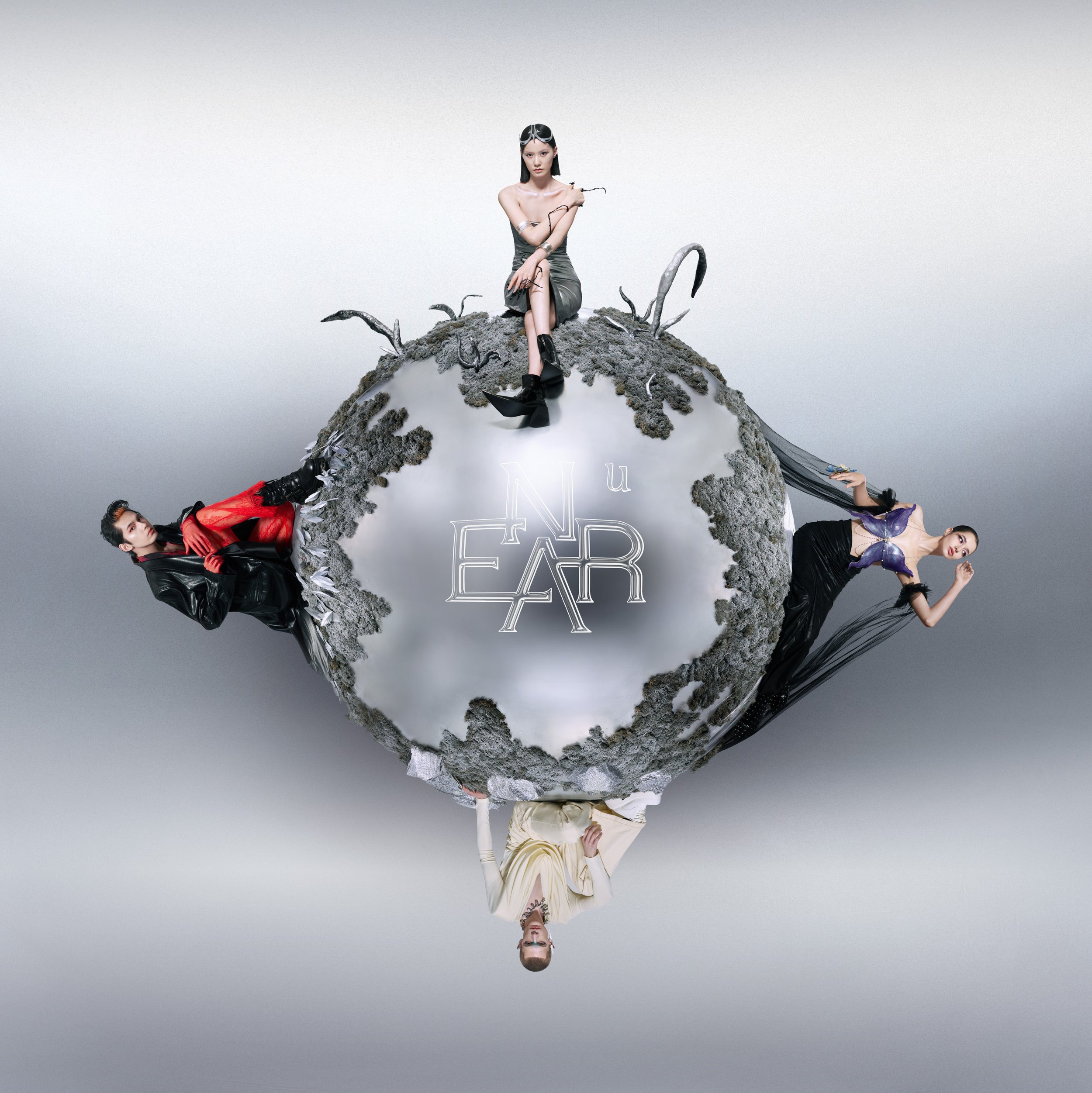 Four women dressed in stylish, fashion-forward outfits are surrounding a small, round, planet-like object with moss and black tentacle-like structures on it. One woman is sitting on top of the sphere, and the others are positioned around the sphere, holding on or leaning against it. The sphere has a reflective surface with the stylized word "NEU EAR" written on it.