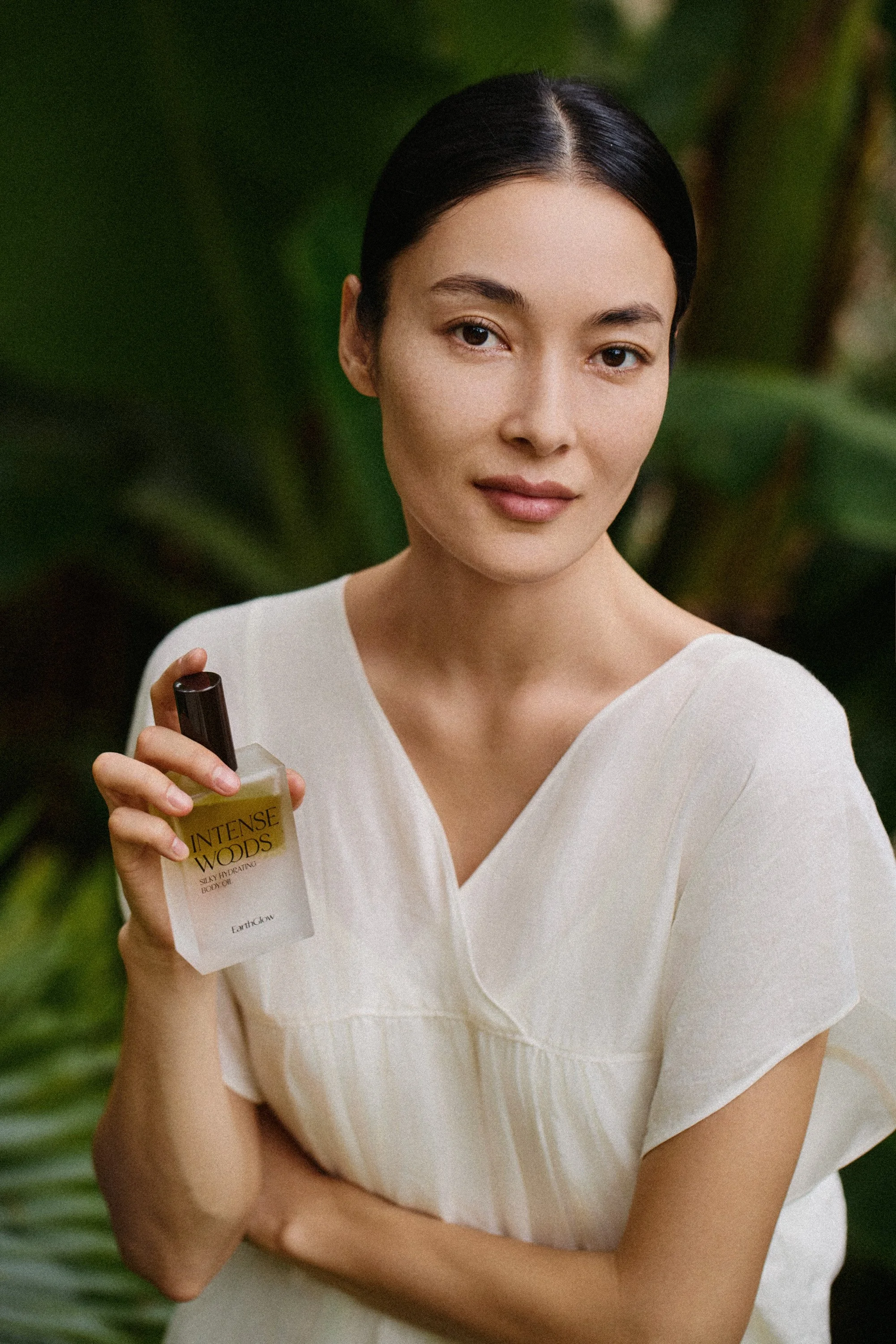 A young woman with dark hair styled back, wearing a white top, holding a bottle of Burt's Bees Intense Woods body lotion, standing outdoors with green foliage in the background.