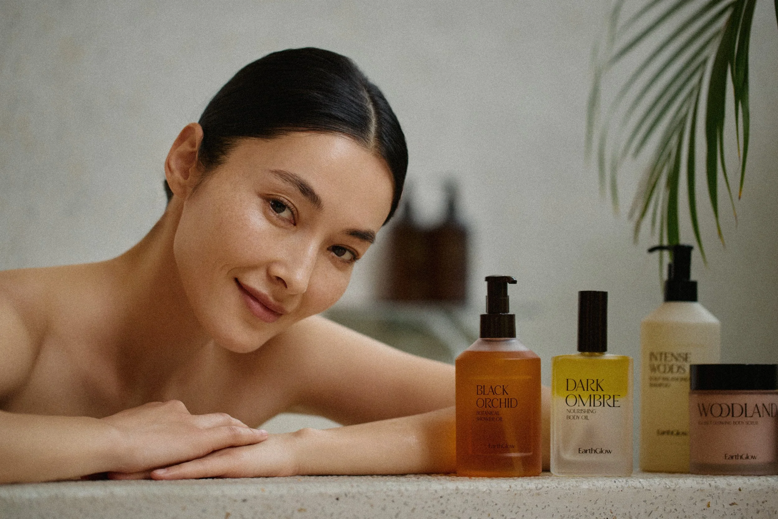 A woman with short dark hair and a gentle smile rests her head on her arms in front of a collection of EarthGlow skincare bottles labeled Black Orchid Botanical Shower Oil, Dark Ombre Nourishing Body Oil, and Woodland Nourishing Body Lotion, with a plant in the background.