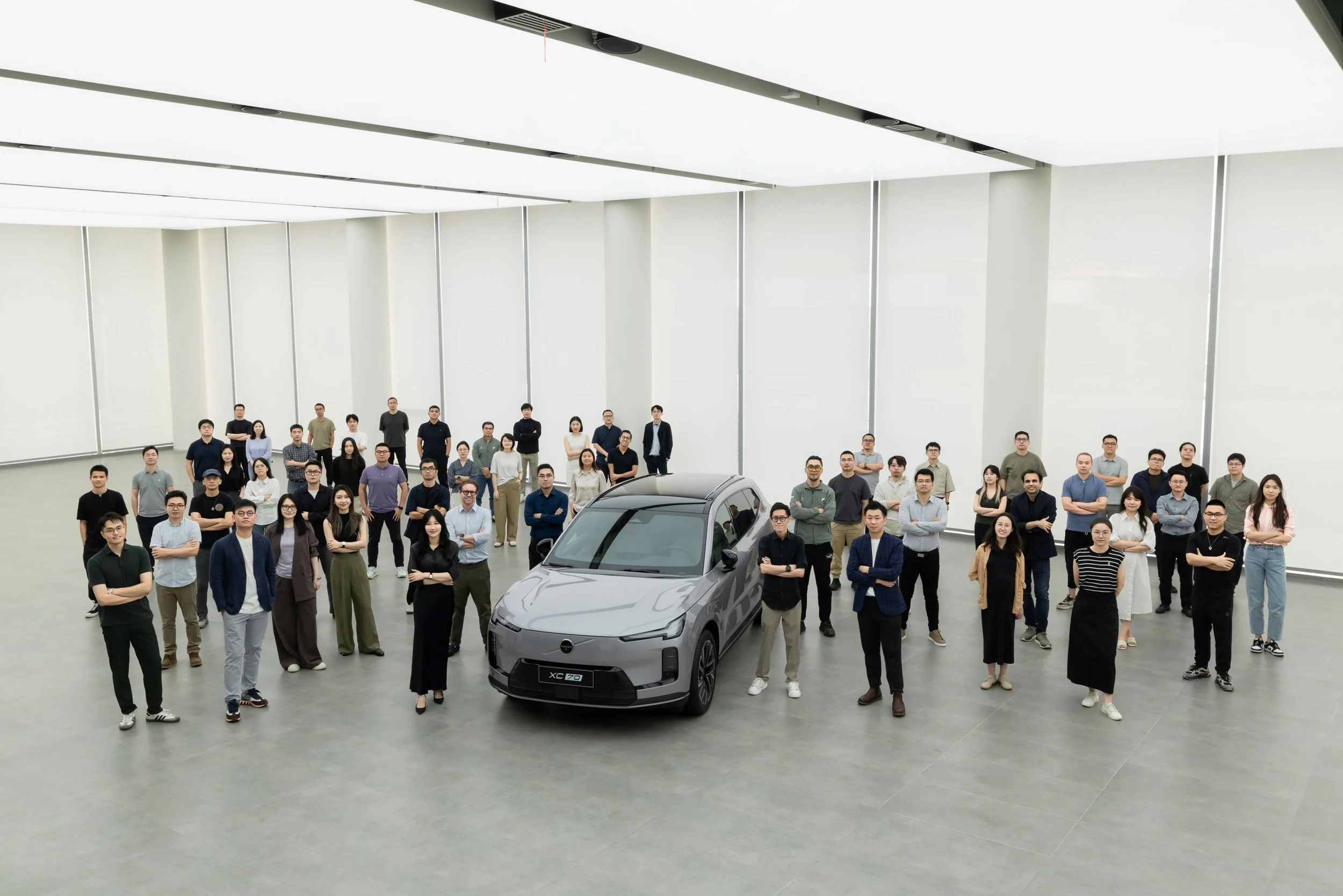 Group of people gathered around a gray car in a bright, spacious, modern indoor setting with high ceilings and large windows.