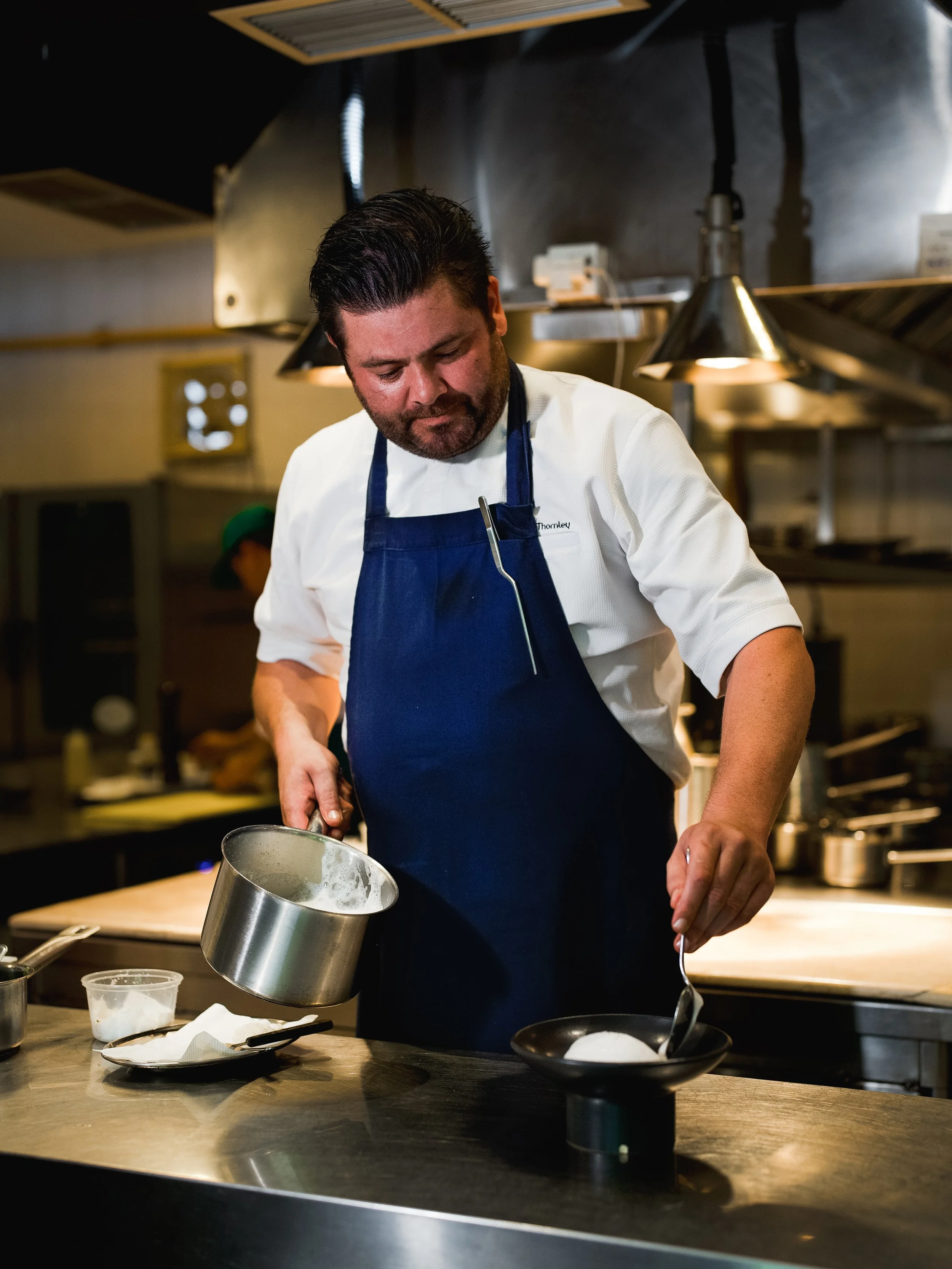 A smiling chef in a black apron and shirt stands in a kitchen, leaning on a countertop.