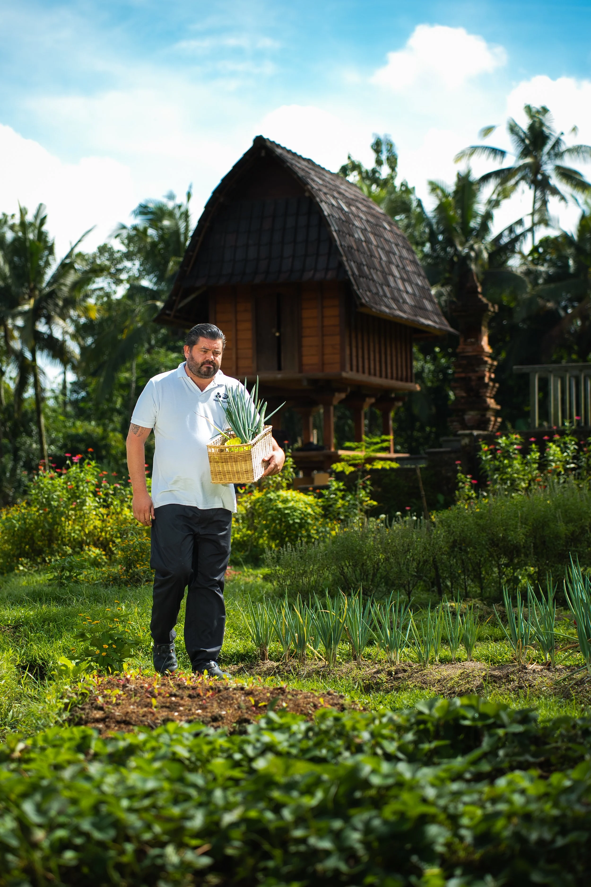 Chef in a restaurant wearing a white chef's uniform and apron, standing in a dining area with tables and chairs in the background.