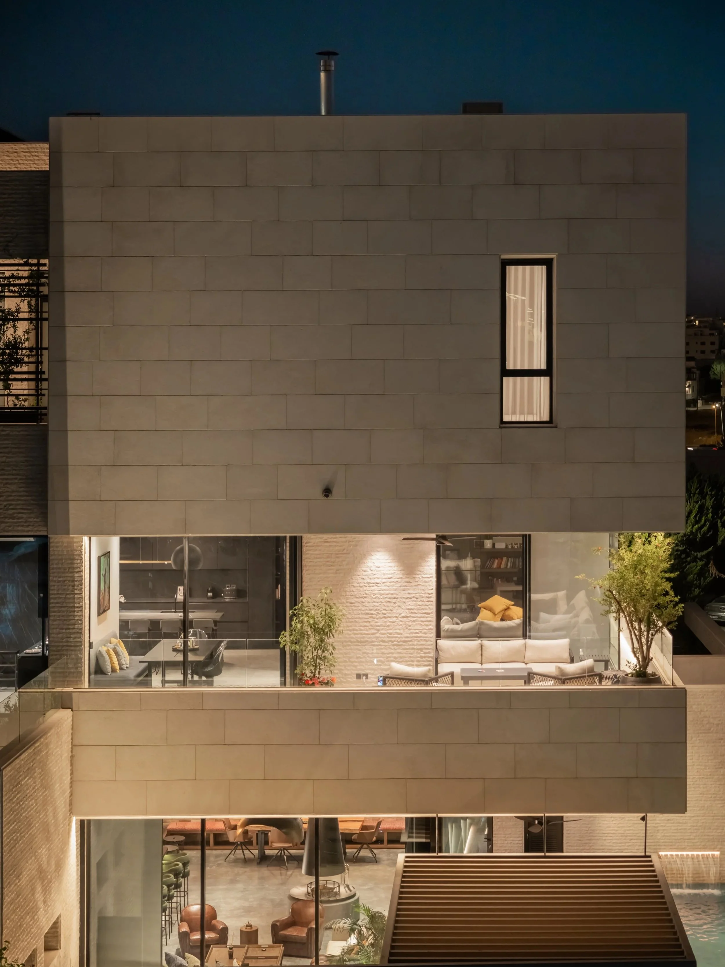 A multi-story modern building with large windows revealing a furnished living space inside. The exterior is constructed of uniform, rectangular, light-colored stone blocks and features a narrow vertical window. The scene appears to be at night with interior lighting illuminating the rooms.