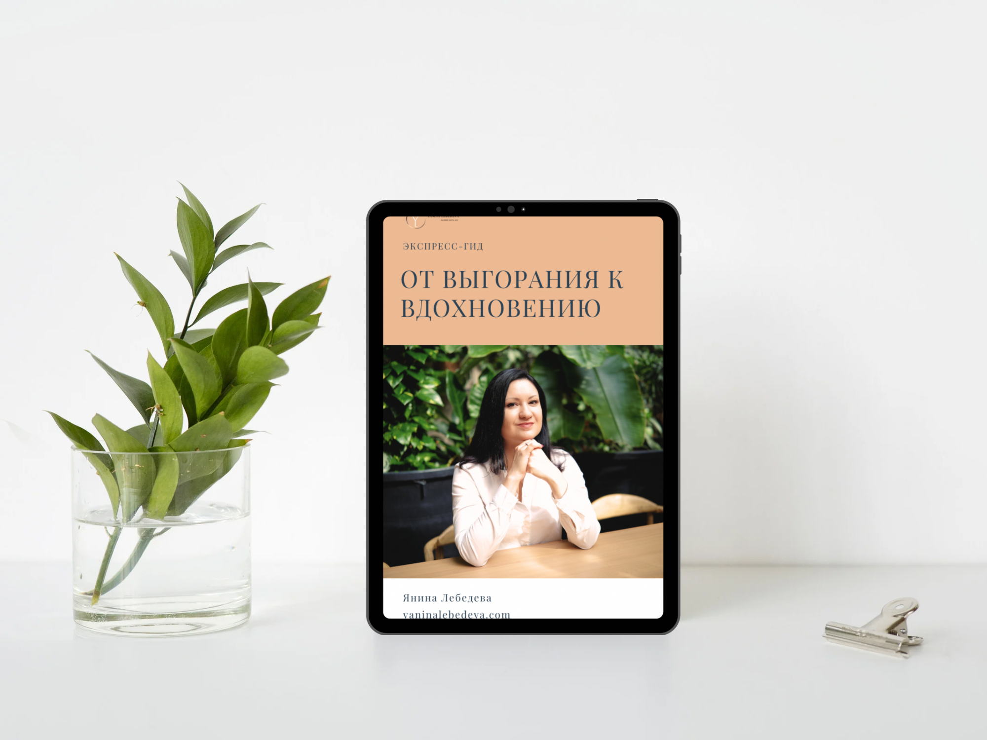 A tablet displaying a book cover in Russian, with a woman sitting at a table in a lush green setting, a vase with green leaves to the left, and a silver clip to the right on a white surface.