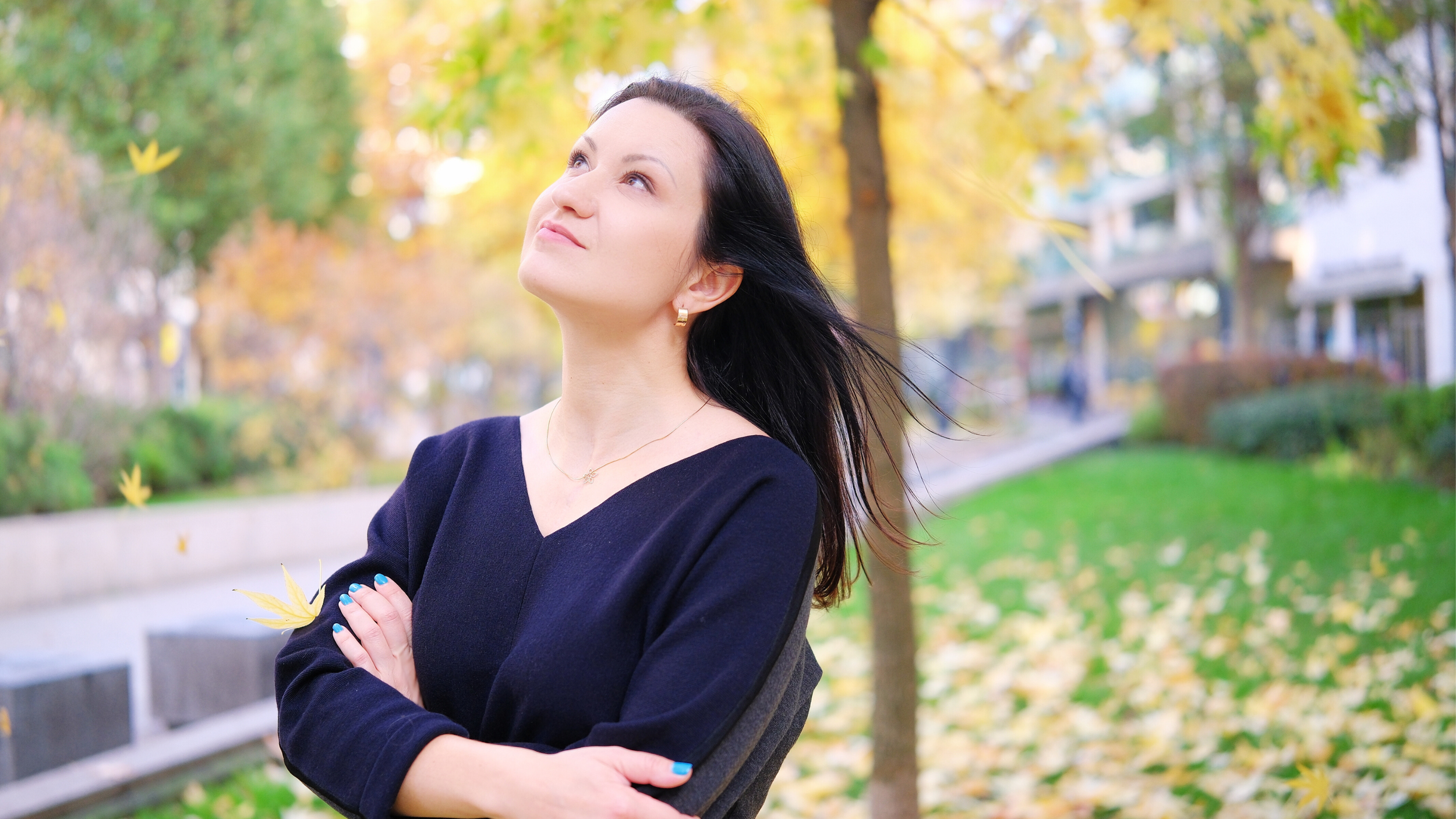 A woman with dark hair and light skin is standing outdoors in a park with trees and fallen leaves, looking up with a contemplative expression.