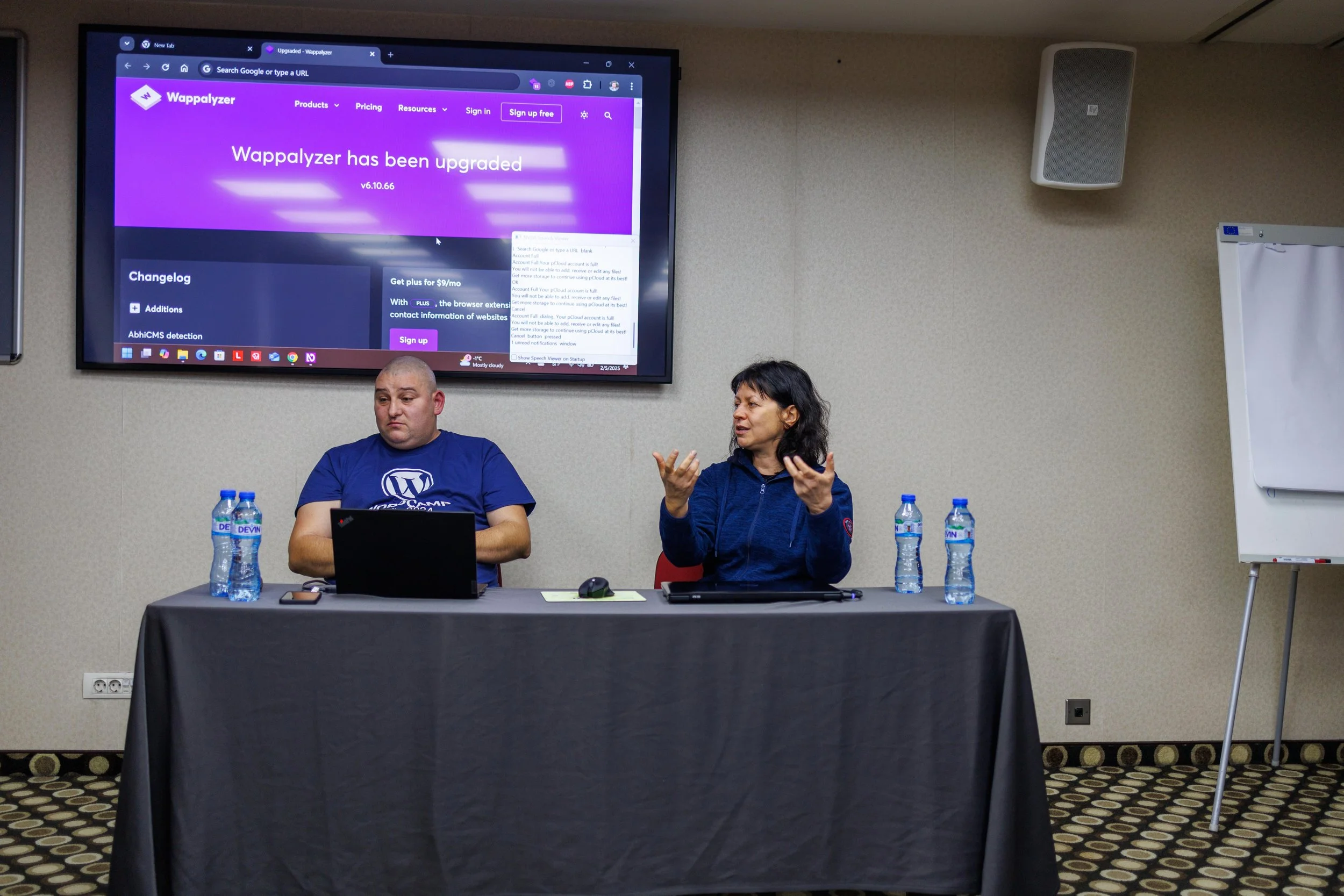 Two people sit at a conference table with three bottles of water each. A man on the left looks at his laptop, while a woman on the right gestures as she speaks. A large screen on the wall behind them displays a website with a purple background.