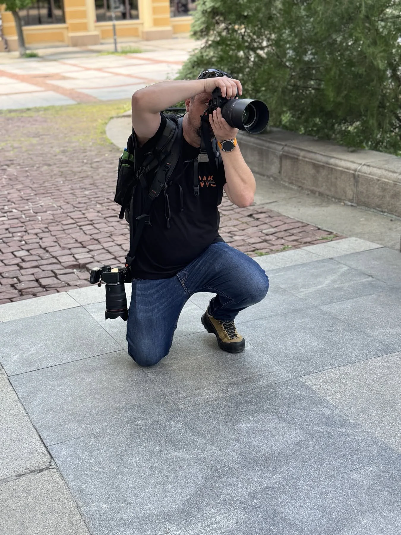 A man kneeling on one knee taking a photograph with a professional camera outdoors on a paved area. He is wearing a black T-shirt, blue jeans, brown hiking shoes, a smartwatch, and has a backpack with a camera lens attached to his belt.
