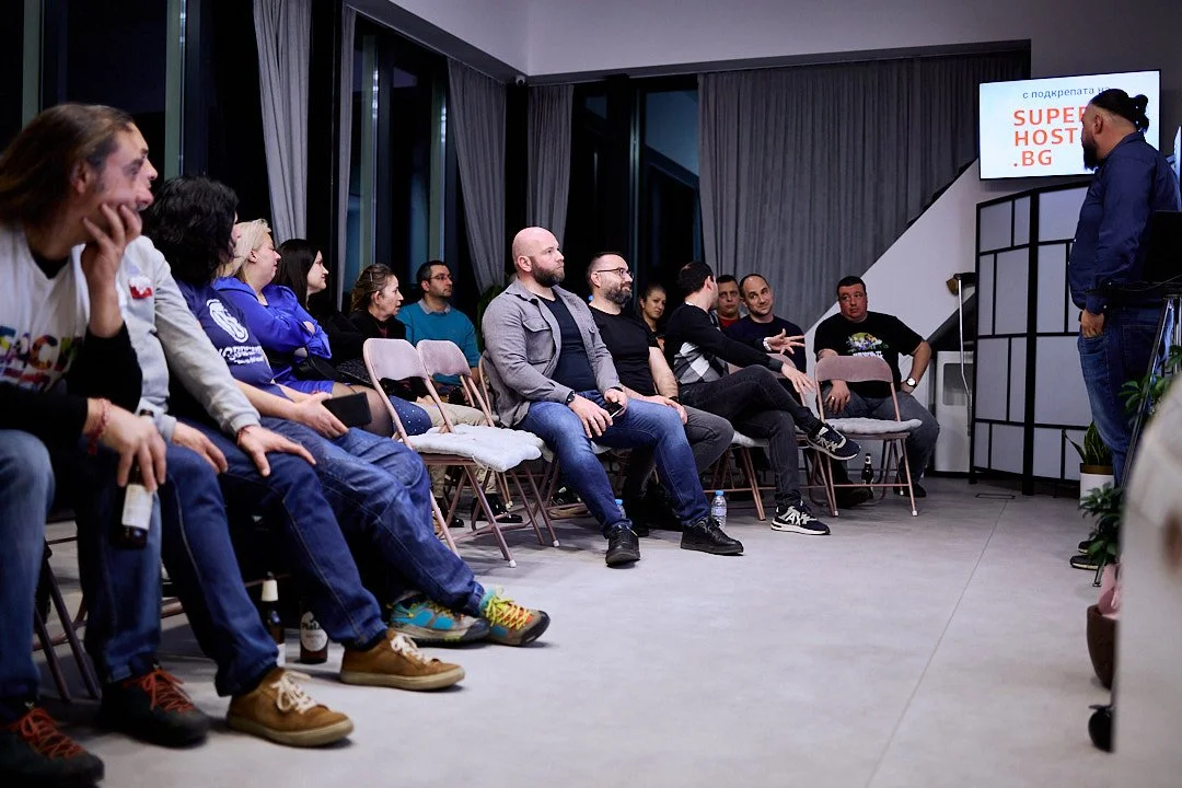A group of people seated in chairs, attending a presentation in a modern room with large windows and gray curtains.